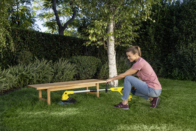 Eine Frau in rosa Shirt und Jeans mäht mit einem gelben Rasentrimmer den Rasen in ihrem Garten.