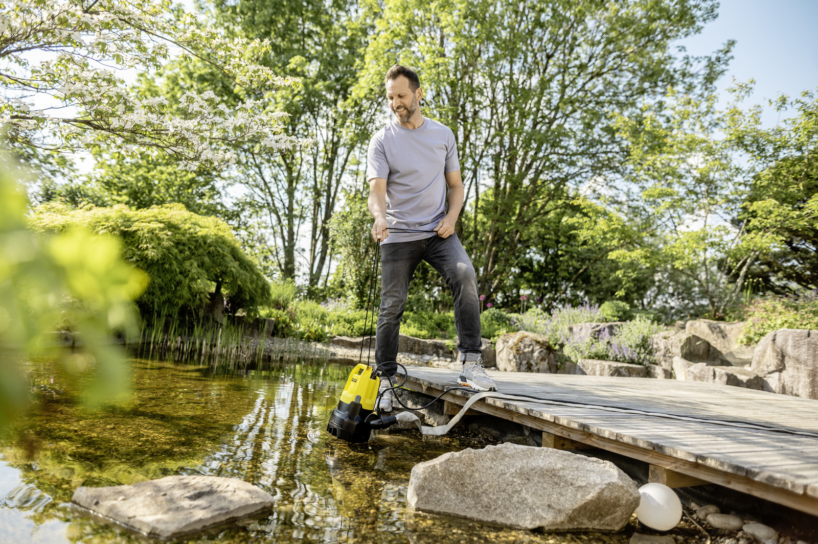 Ein Mann steht auf einem Holzsteg an einem Gartenteich und hält eine gelbe Schmutzwasserpumpe.
