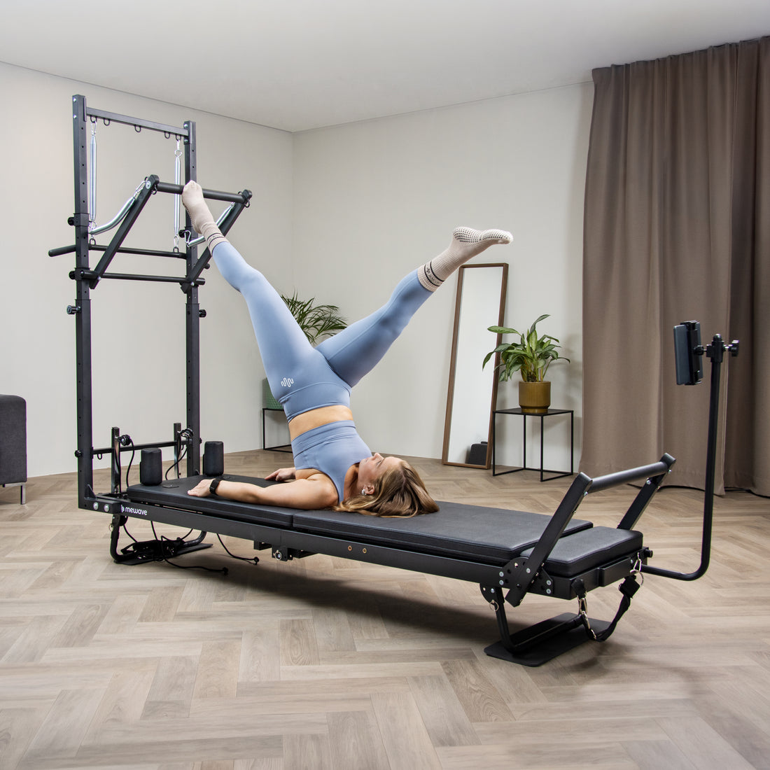Una mujer con ropa deportiva azul hace ejercicio en una máquina reformer negra en una habitación luminosa.