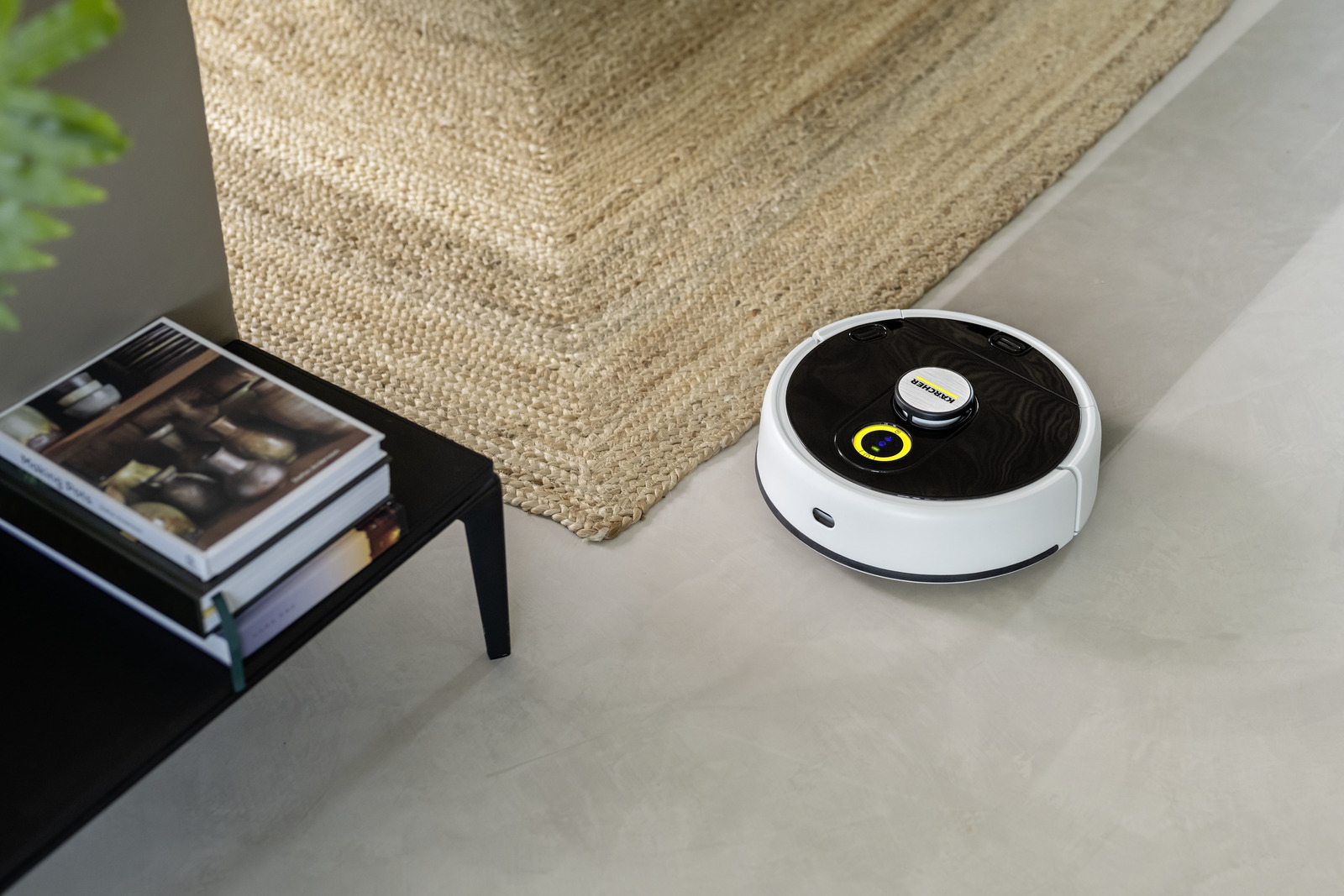 Un robot aspirapolvere bianco su un pavimento in cemento vicino a un tappeto e un tavolo con libri.