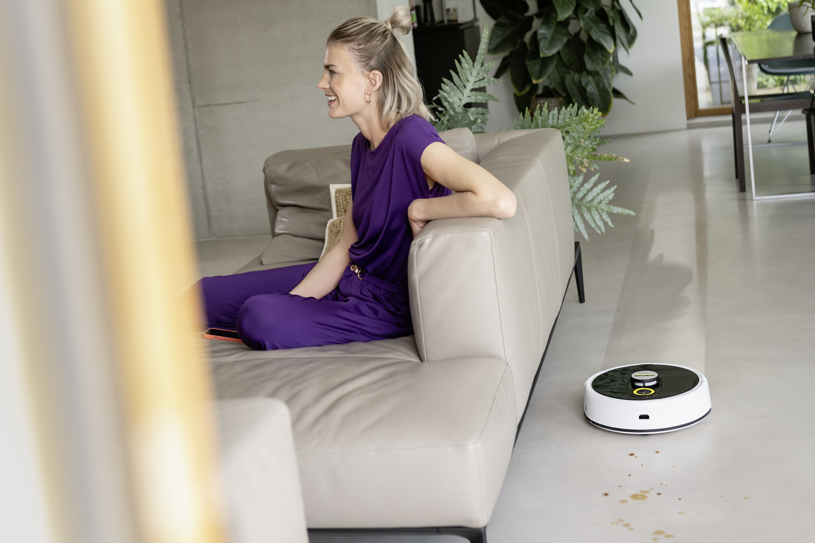 Una donna con abiti viola siede su un divano mentre un robot aspirapolvere pulisce il pavimento.