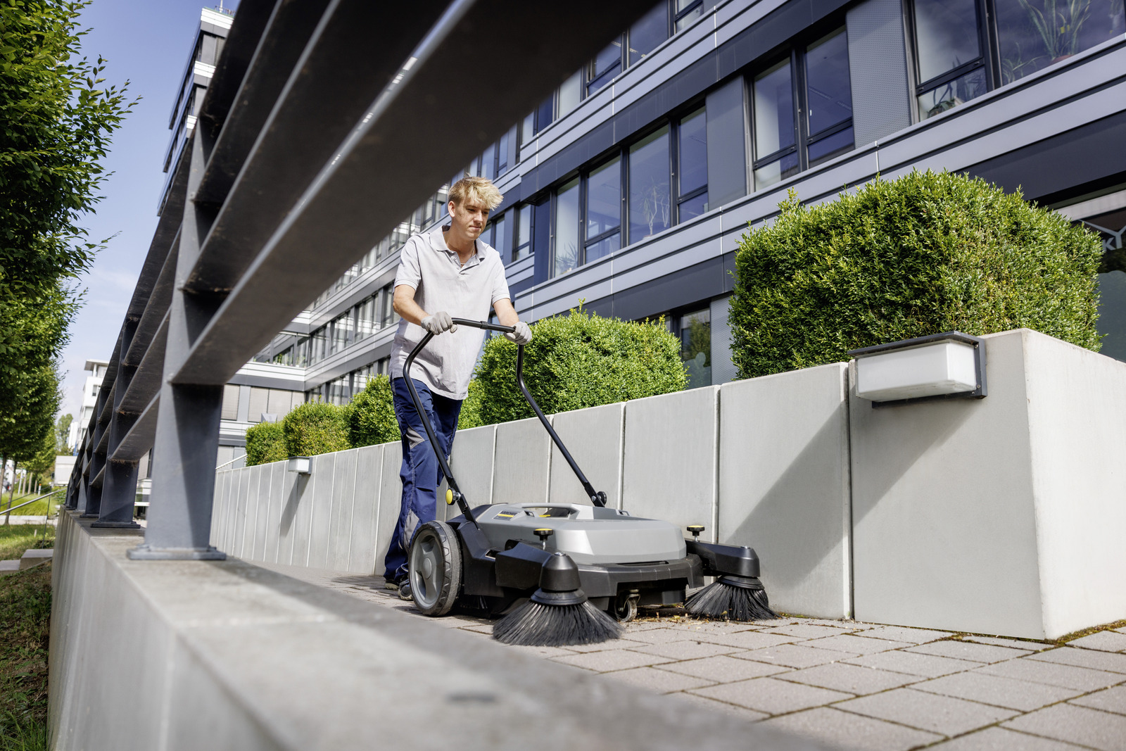 Ein Arbeiter bedient eine Kehrmaschine auf einem Gehweg vor einem modernen Bürogebäude.