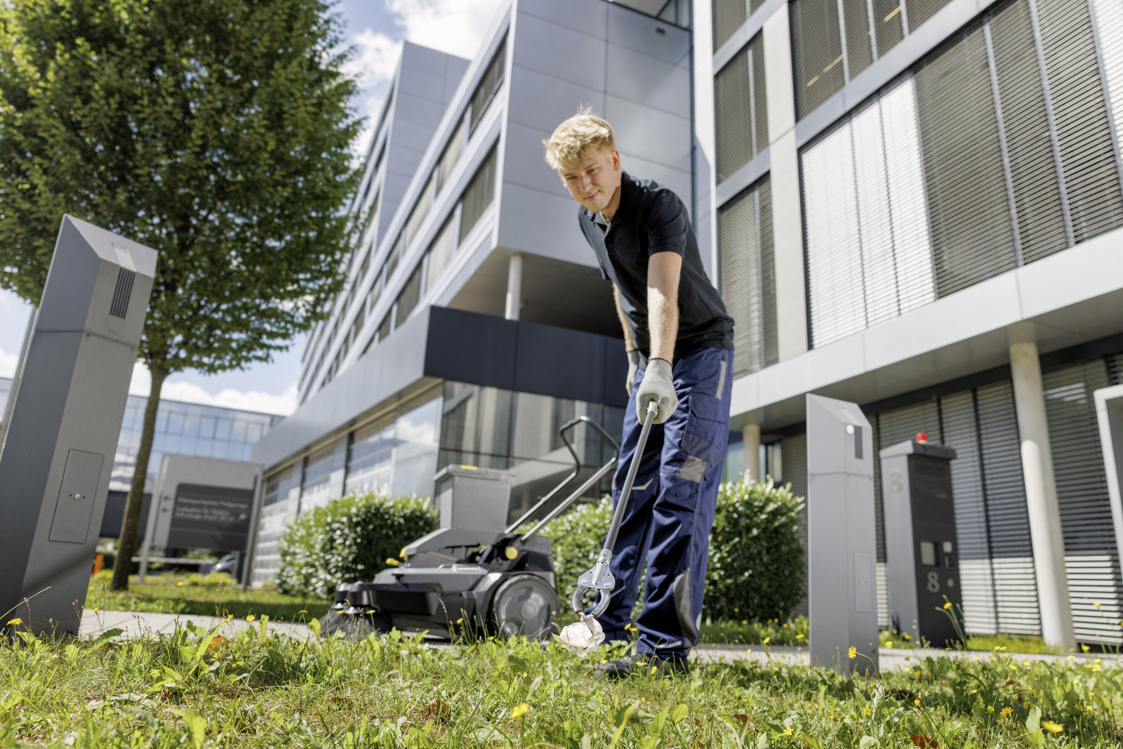 Ein Arbeiter in blauer Hose sammelt mit einer Zange Müll vom Gras vor einem modernen Gebäude auf.