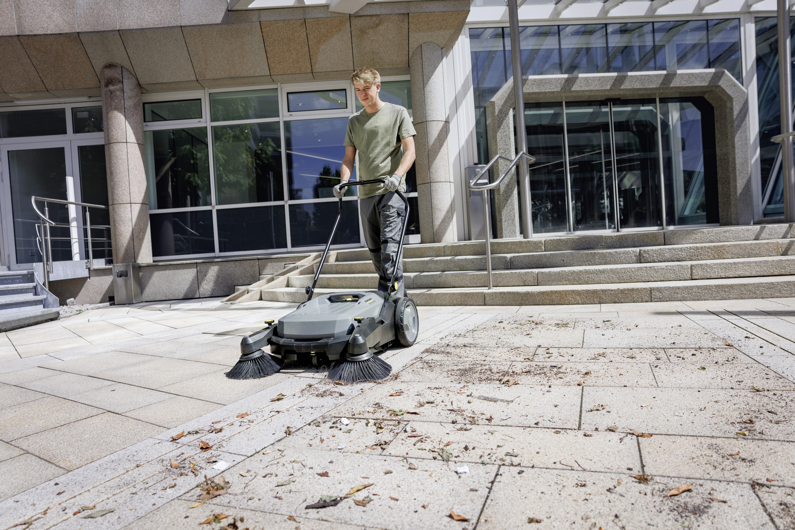 Ein Arbeiter reinigt mit einer Kehrmaschine den gepflasterten Vorplatz eines modernen Bürogebäudes.