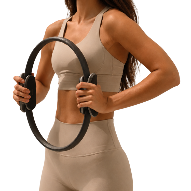 Una mujer con ropa deportiva beige sostiene un anillo de pilates negro frente a su abdomen.