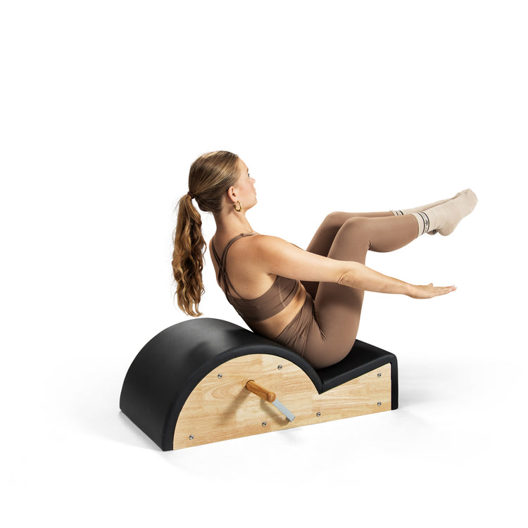 Una mujer en ropa deportiva marrón hace ejercicio en un arco de Pilates negro y de madera.
