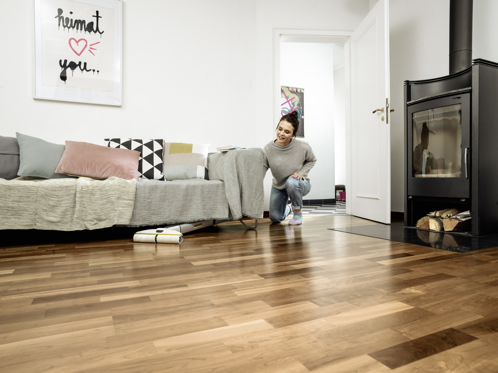 Een vrouw knielt op een houten vloer bij een bank terwijl een vloerreiniger eronder schoonmaakt.