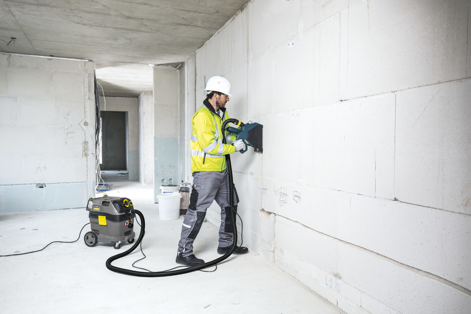 Un lavoratore con casco bianco e giacca gialla usa un elettroutensile con tubo flessibile su un muro bianco.
