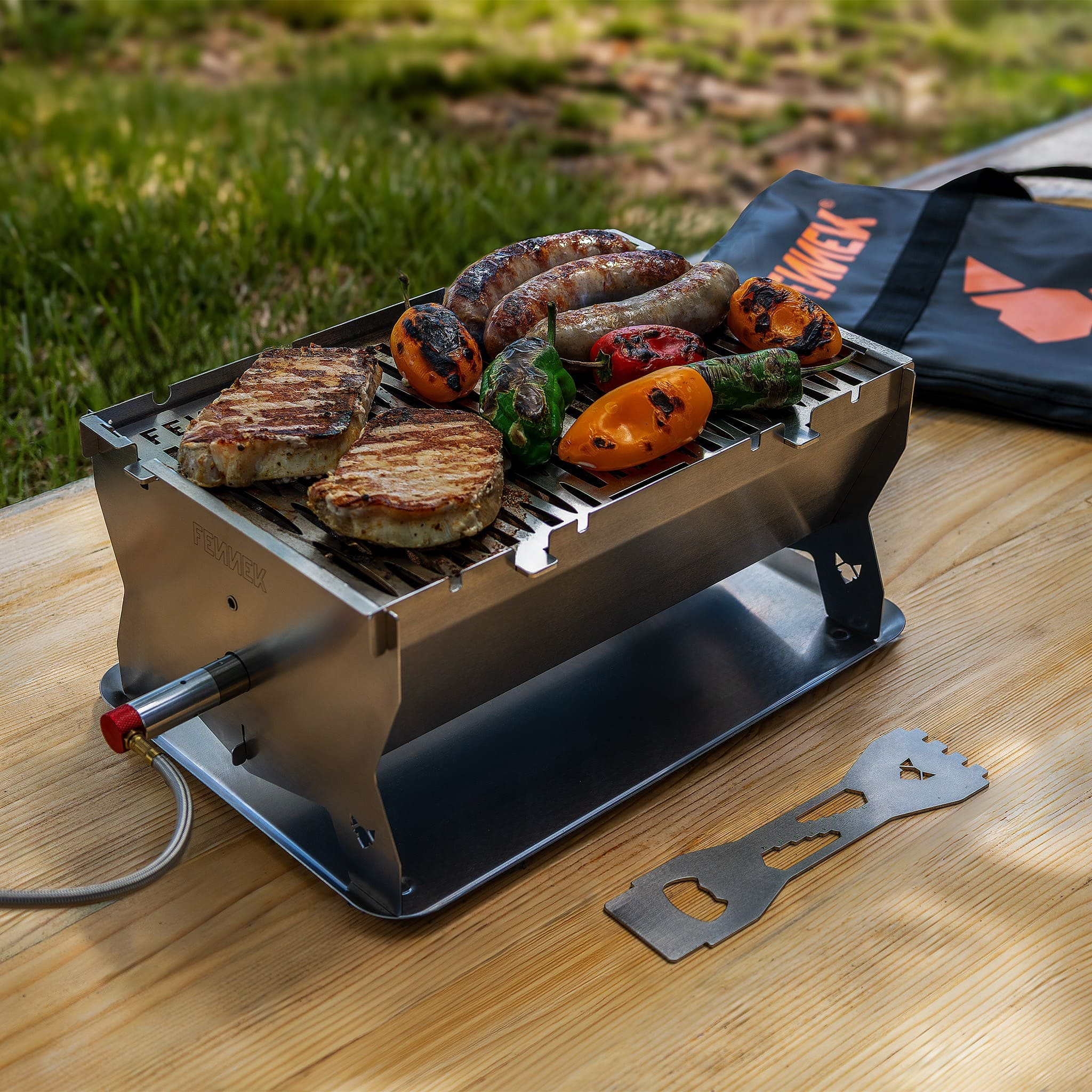 Ein Tischgrill auf Holz kocht Fleisch und Gemüse, mit einem Werkzeug daneben und einer schwarzen Tasche im Hintergrund.