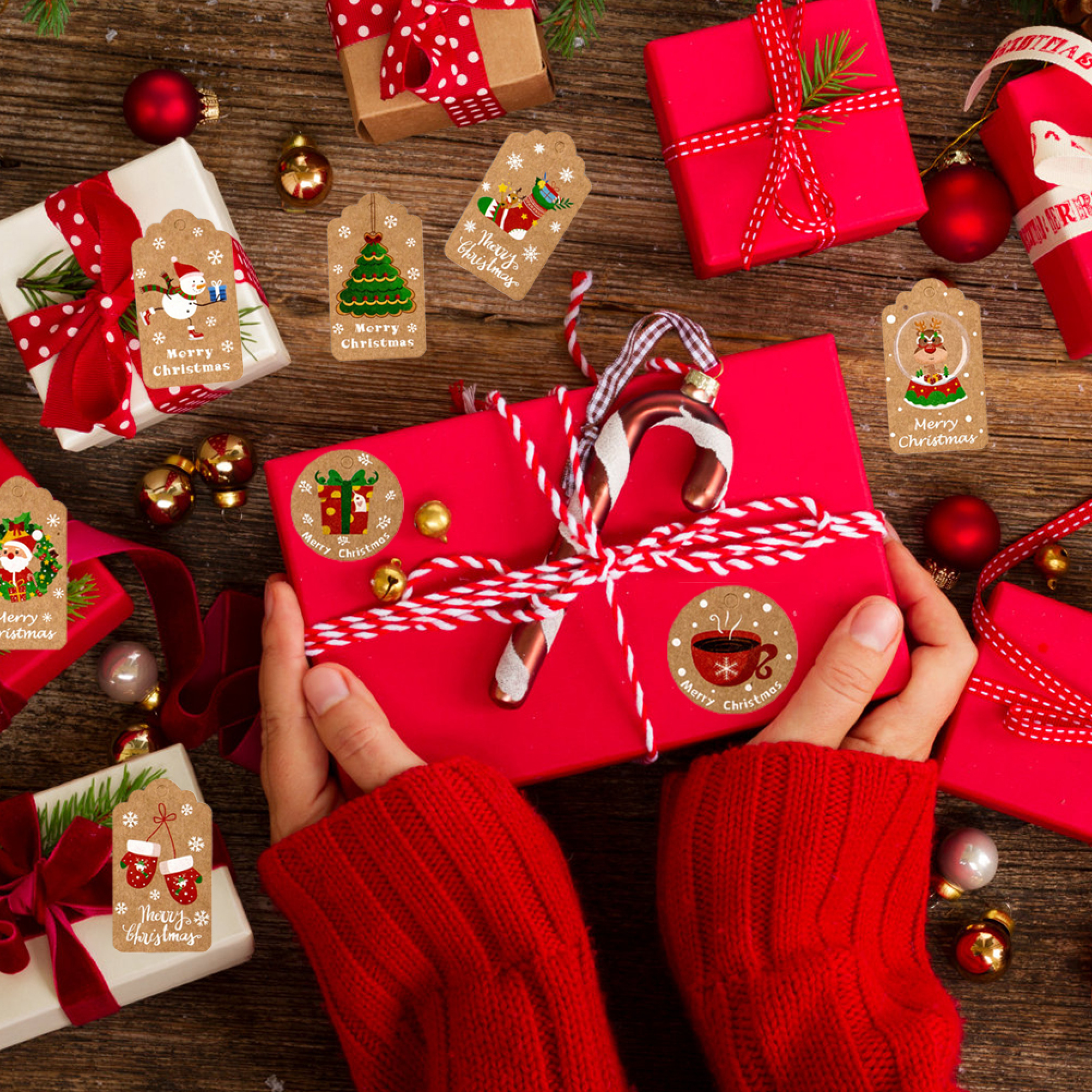 Person in rotem Pullovern hält ein großes rotes Weihnachtsgeschenk umgeben von Geschenken und Anhängern.