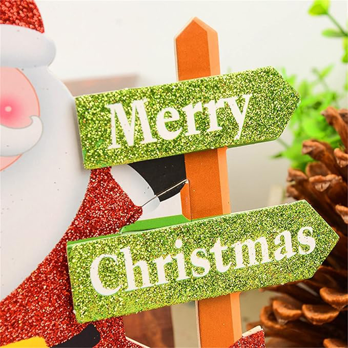 Ein festliches Holzschild mit grünen Glitzerpfeilen mit der Aufschrift Frohe Weihnachten, neben einer Figur und einem Tannenzapfen.
