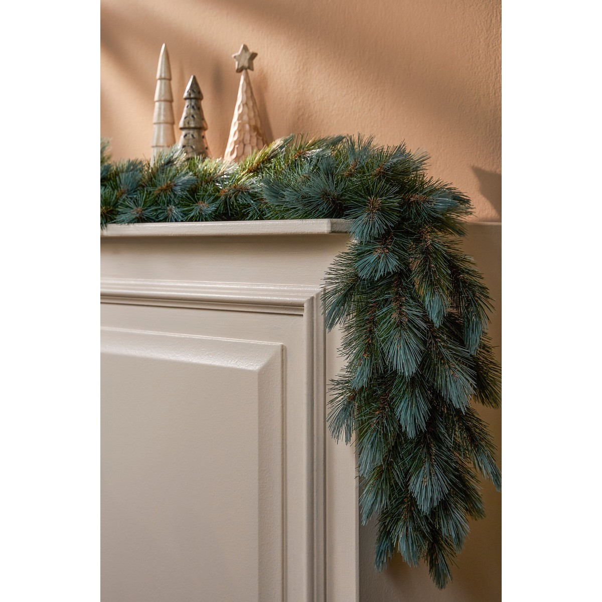 Een dennenguirlande hangt over een beige schoorsteenmantel, met drie kleine decoratieve houten bomen erbovenop.