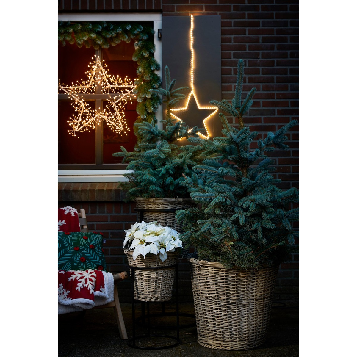 Kerstdecoratie voor een bakstenen muur met kerstbomen in manden, verlichte sterren en een stoel.