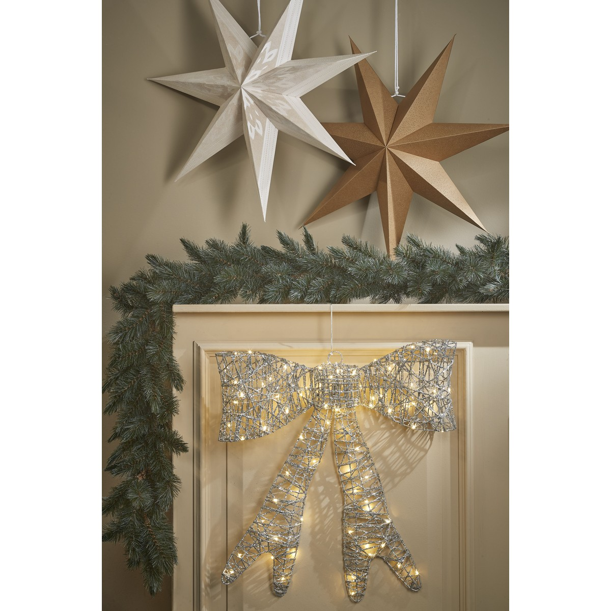 Kerstdecoratie met twee hangende sterren, een slingers en een verlichte zilveren strik aan een muur.