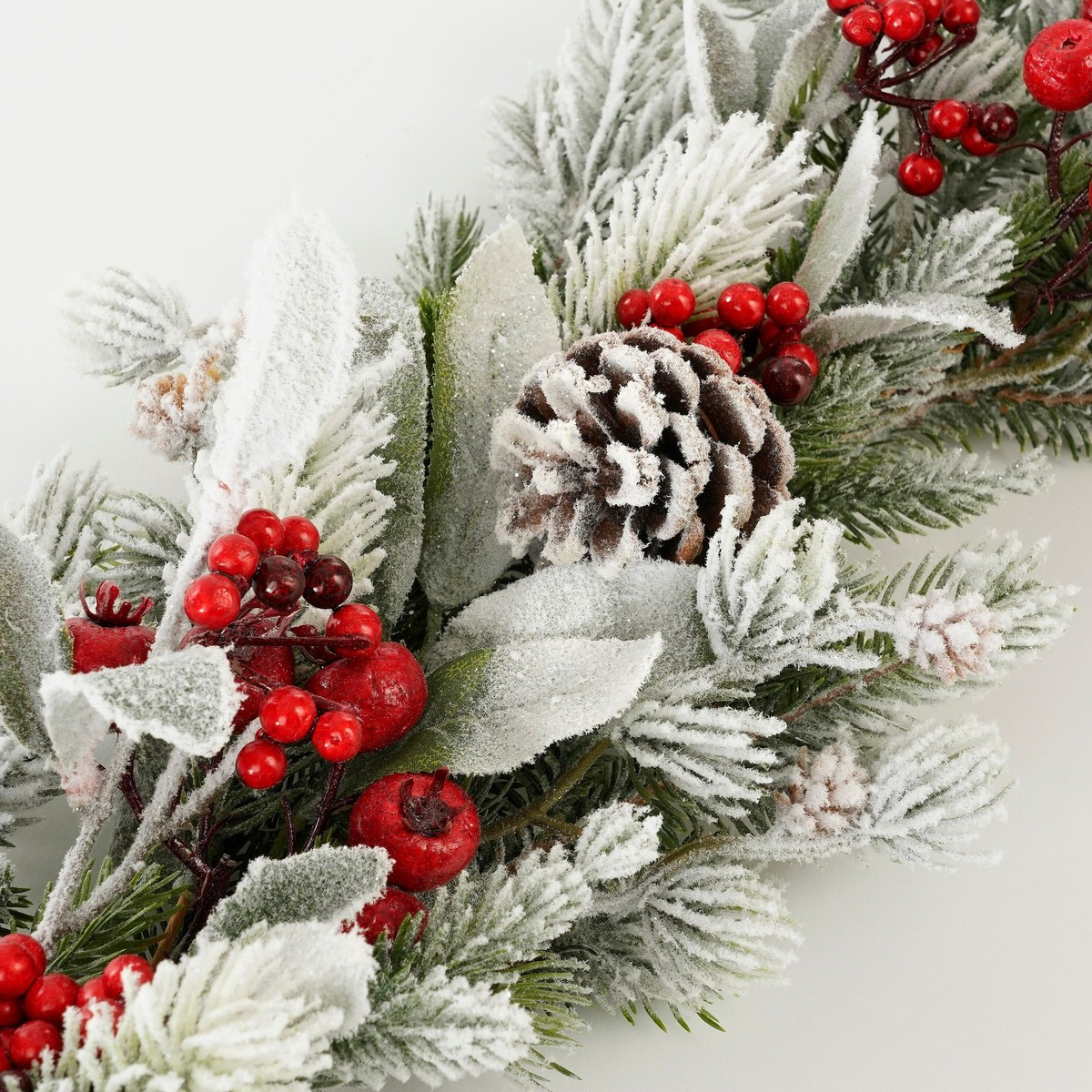 Guirlande de Noël artificielle givrée avec pommes de pin, feuilles vertes et baies rouges sur fond blanc.