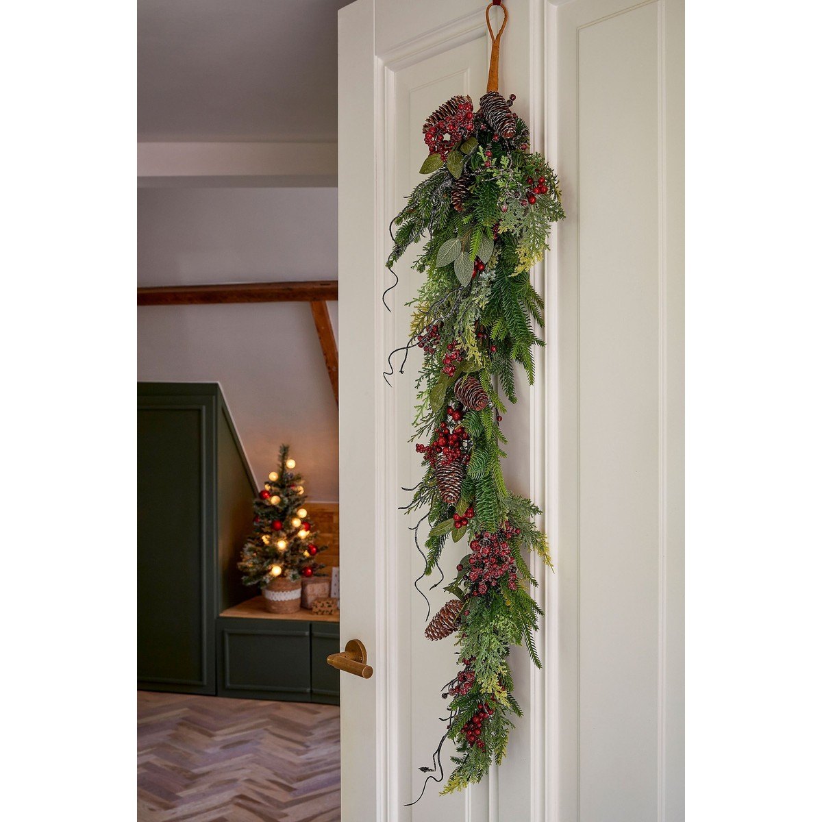 Een kerstslinger van dennengroen, dennenappels en rode bessen hangt aan een witte deur, met een klein verlicht boompje op de achtergrond.