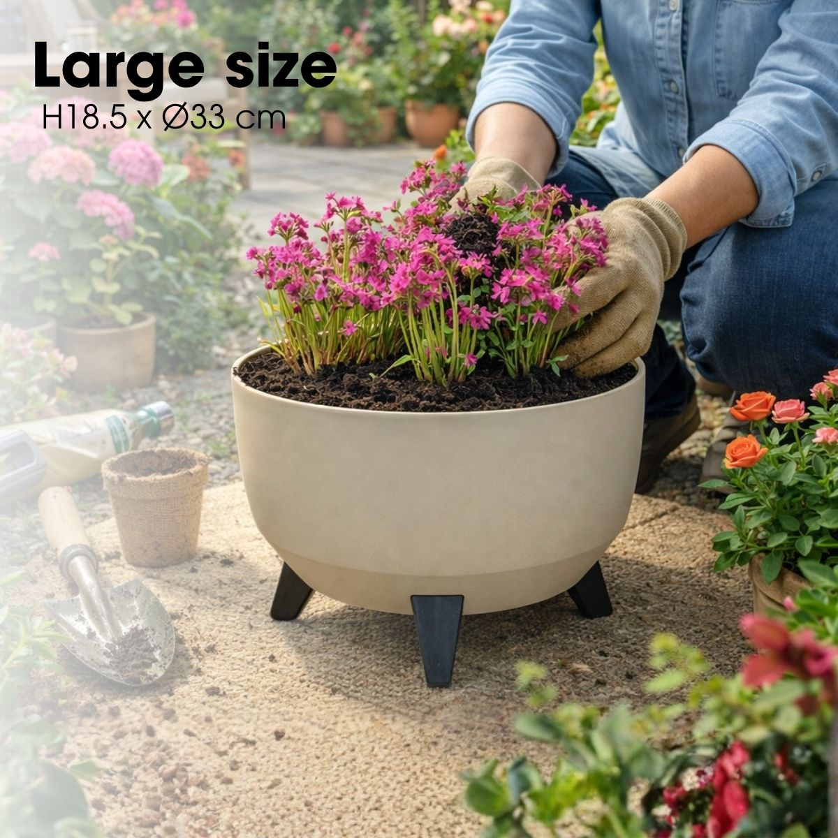 Une personne avec des gants plante des fleurs roses dans un grand pot beige à trois pieds.