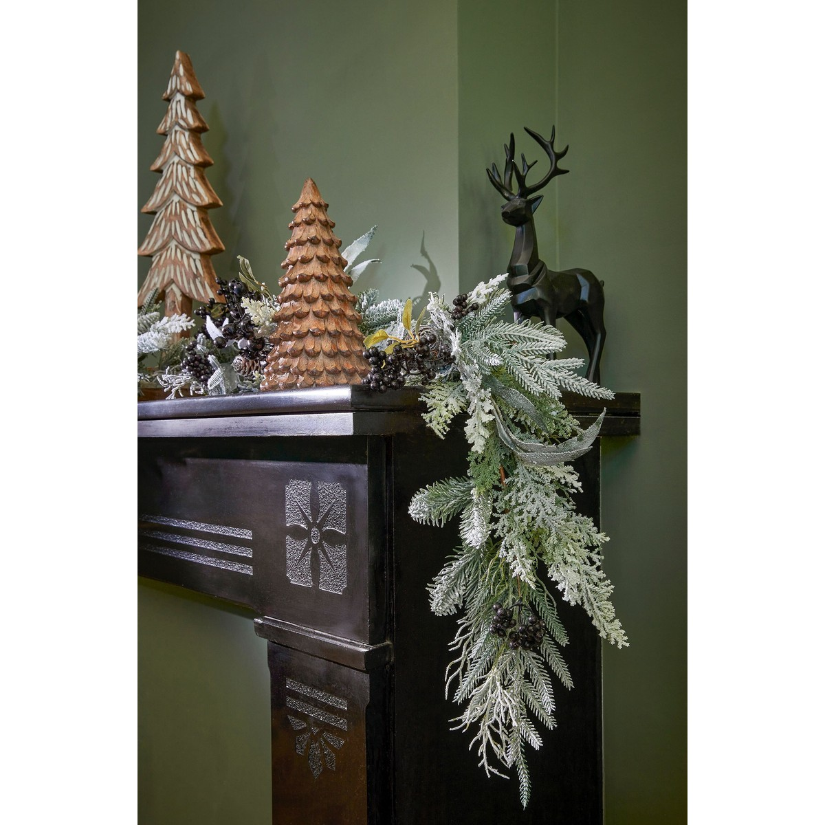 Kerstopenhaarddecoratie met houten dennenbomen, een zwart hert en een besneeuwde denneguirlande tegen een groene muur.