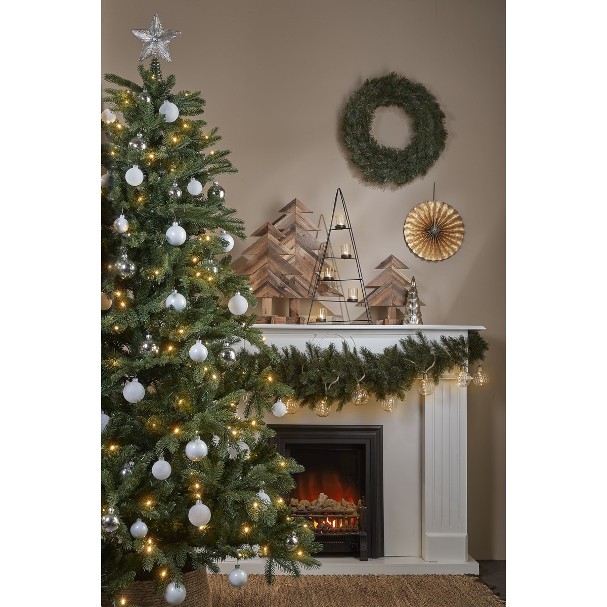 Versierde kerstboom naast open haard met slingers, houten bomen en krans.