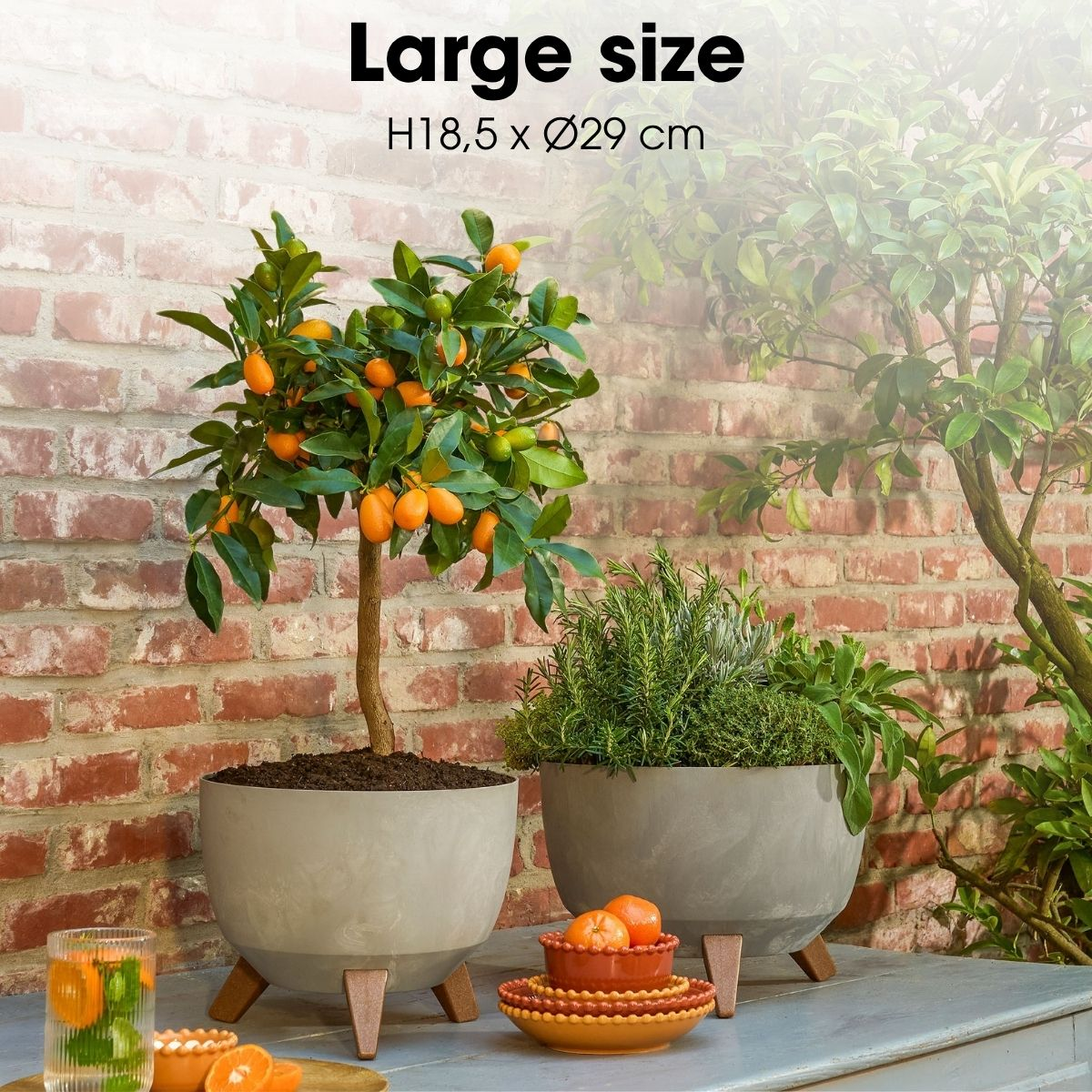 Deux pots ronds gris avec des plantes sur pieds en bois devant un mur de briques.
