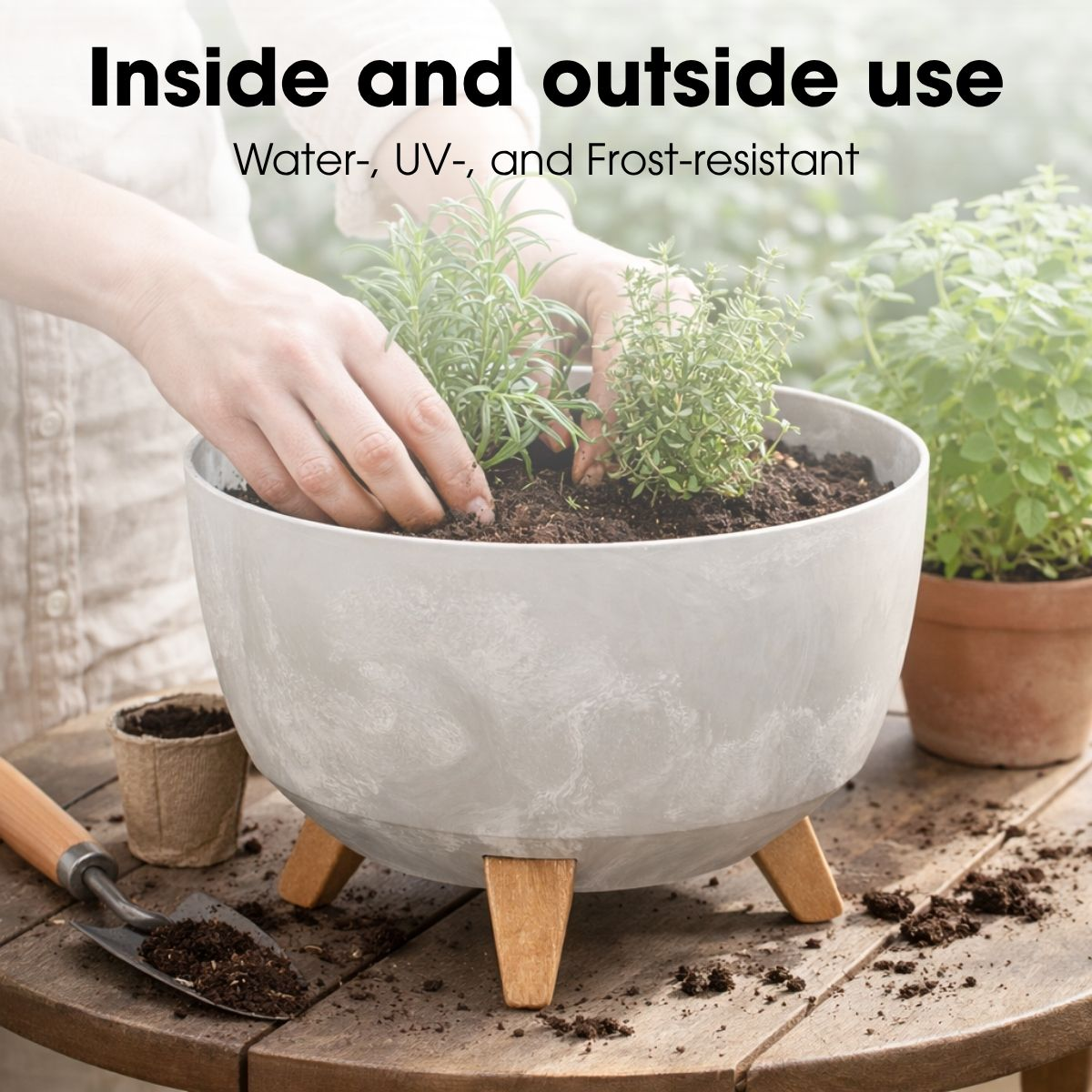 Une personne plante des herbes dans un pot gris à pieds en bois sur une table, avec de la terre et une pelle.