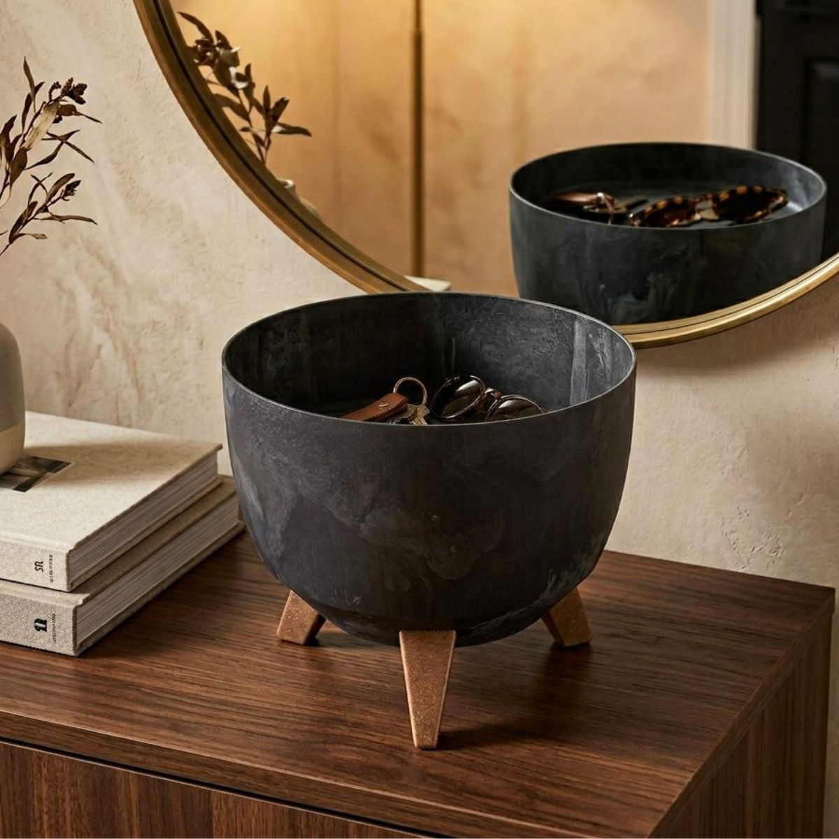 Un bol noir sur trois pieds en bois repose sur une table en bois, avec des clés et un miroir rond.