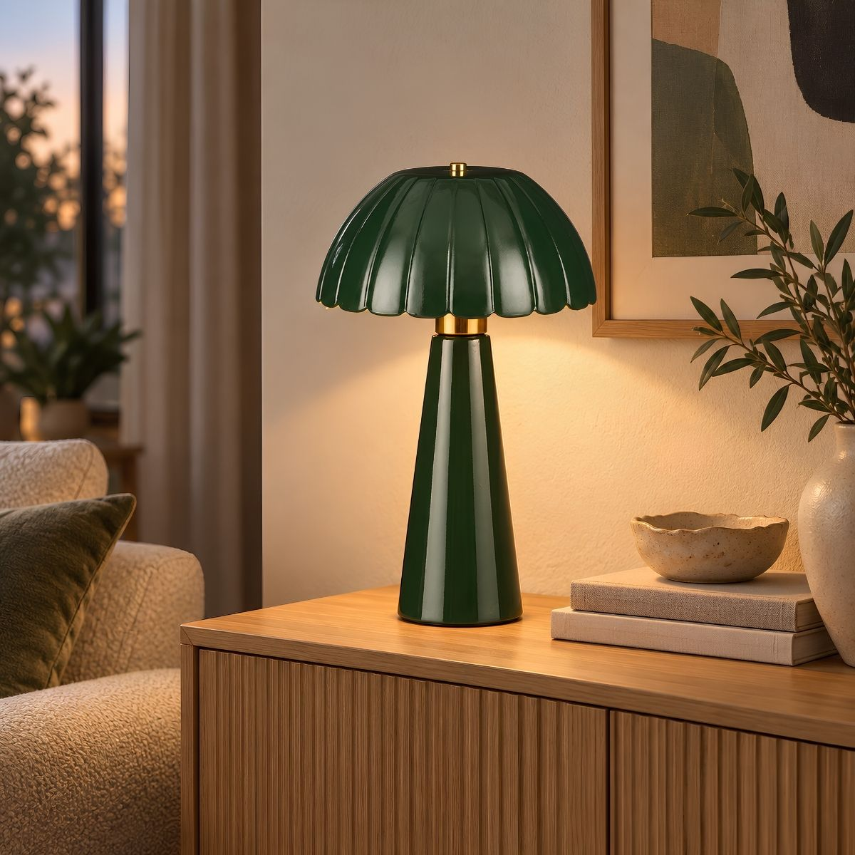 Eine grüne Tischlampe mit geschwungenem Schirm steht auf einer Holzkommode, daneben Bücher und eine Vase mit Zweigen.
