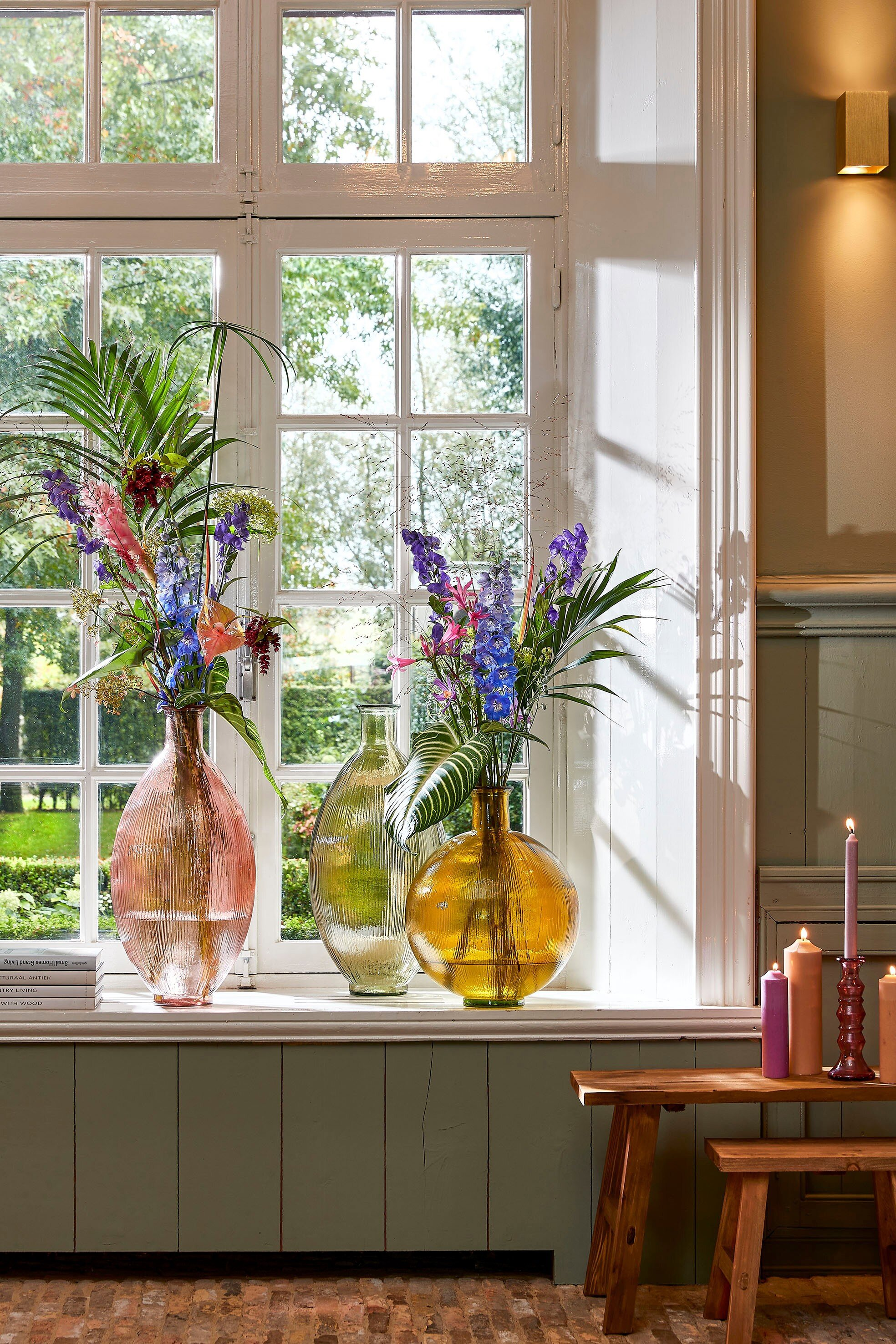Drei bunte Glasvasen mit Blumen stehen auf einer Fensterbank, daneben brennen Kerzen.