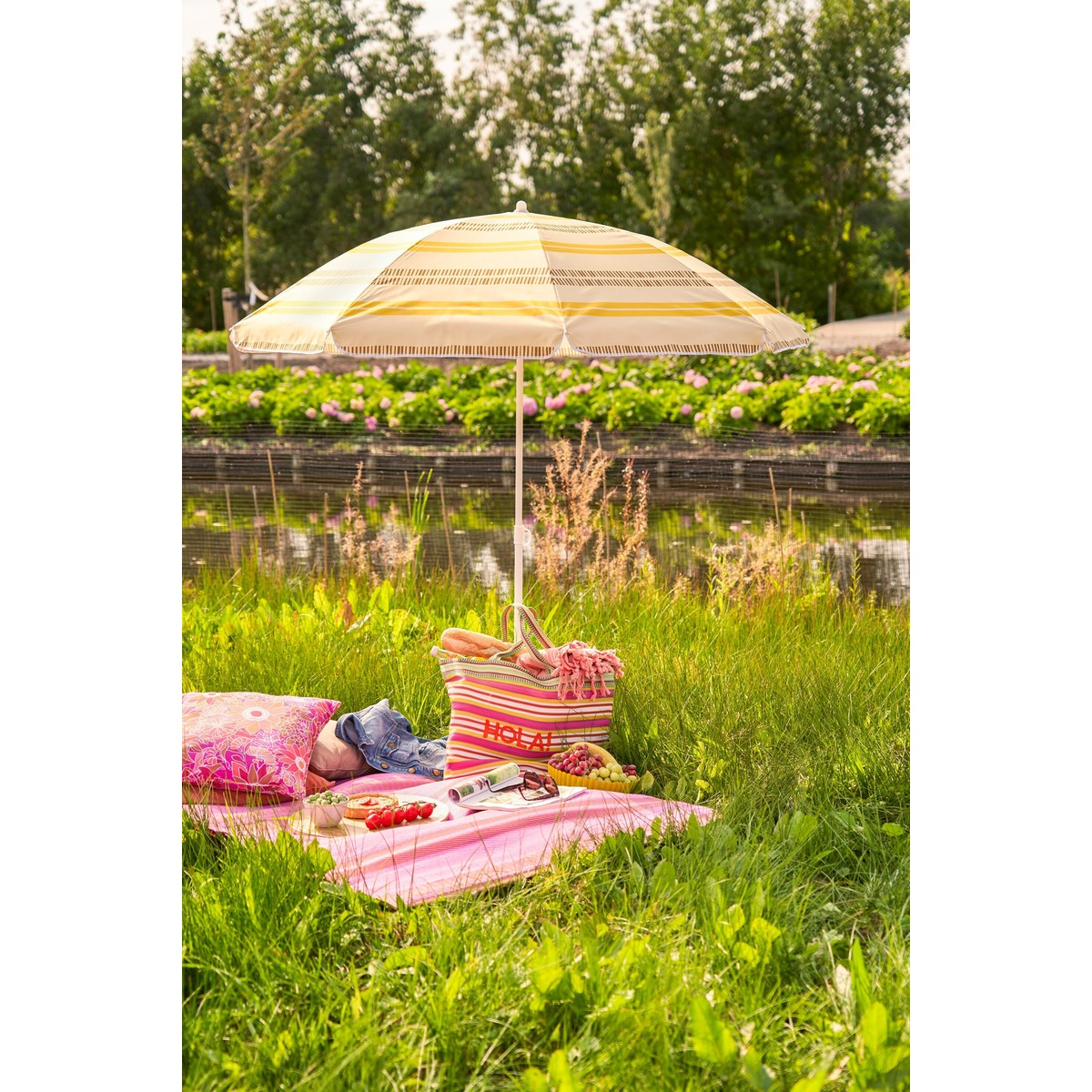 Ein gestreifter Sonnenschirm deckt eine Picknickdecke mit Essen und einer Tasche auf einer Wiese am Wasser.