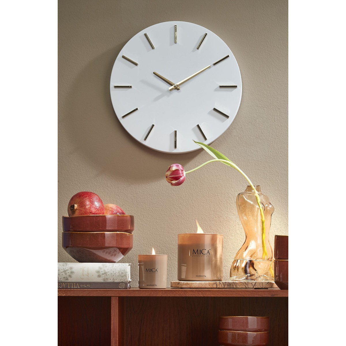 Eine weiße Wanduhr mit goldenen Zeigern hängt über einem Holzregal mit Schalen, brennenden Kerzen und einer Blume.