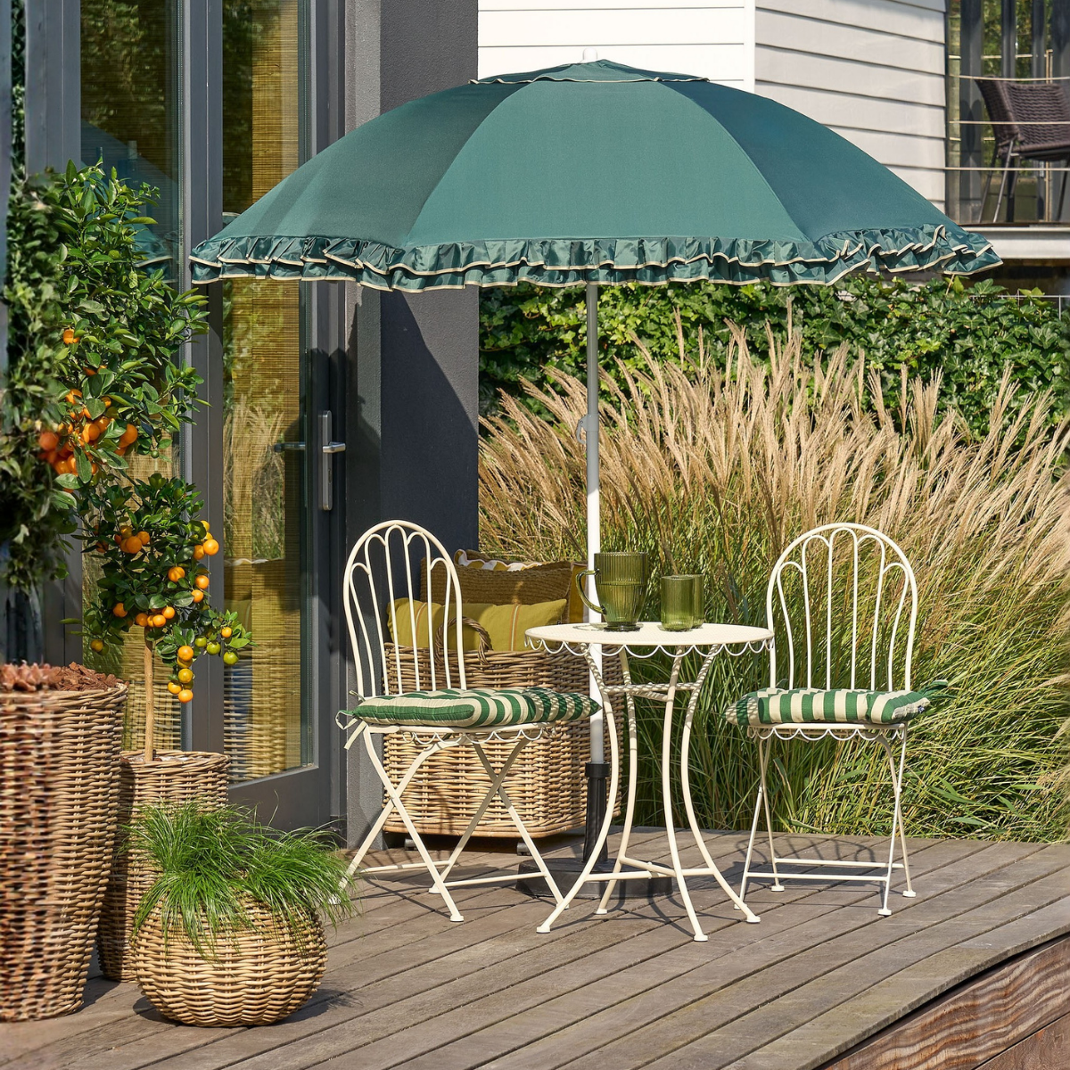 Ein kleiner weißer Metalltisch mit zwei Stühlen und einem grünen Sonnenschirm auf einer Holzterrasse.