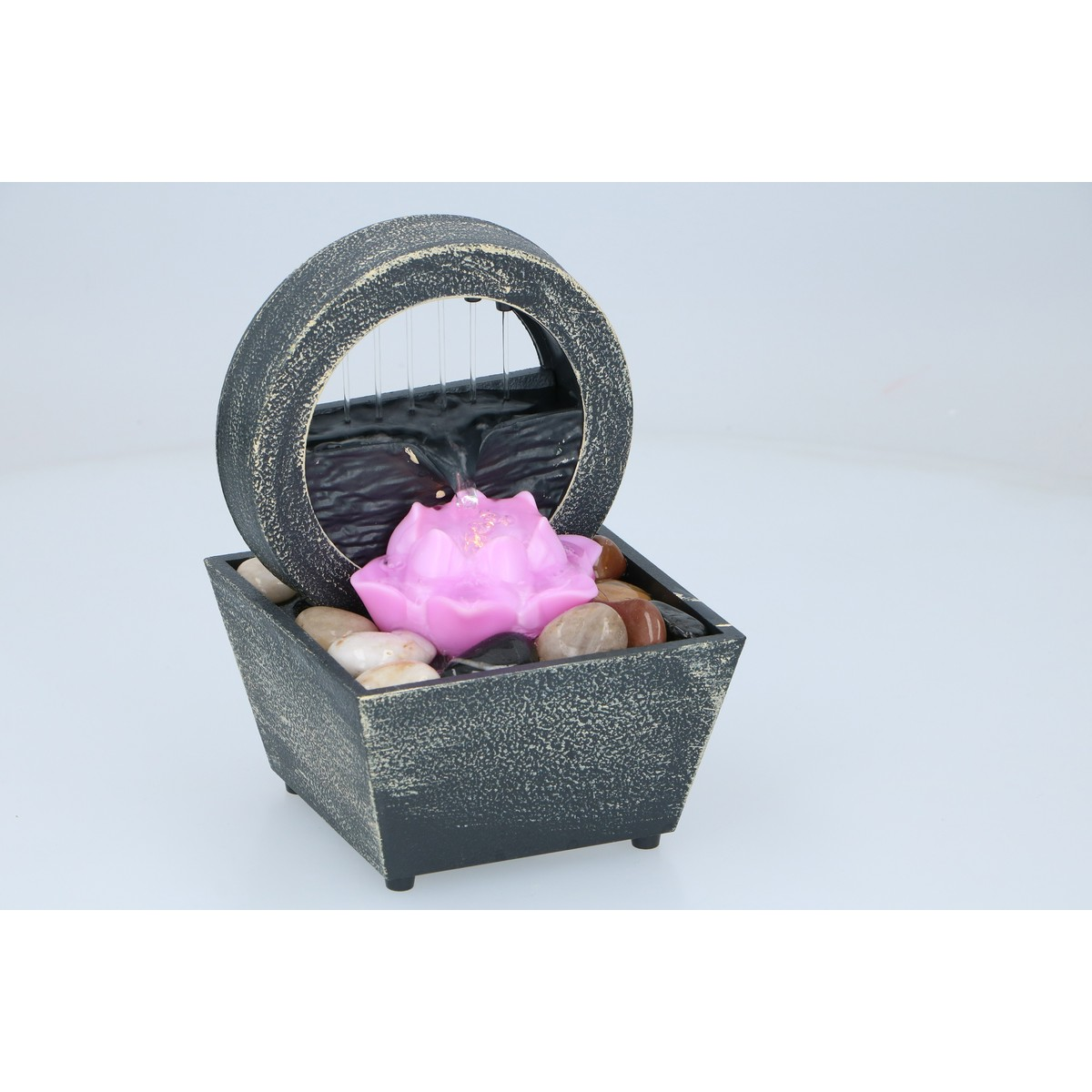 Een decoratieve binnenwaterfontein met een cirkelvormige ring, een roze lotusbloem en gladde keien in een donkere bak.