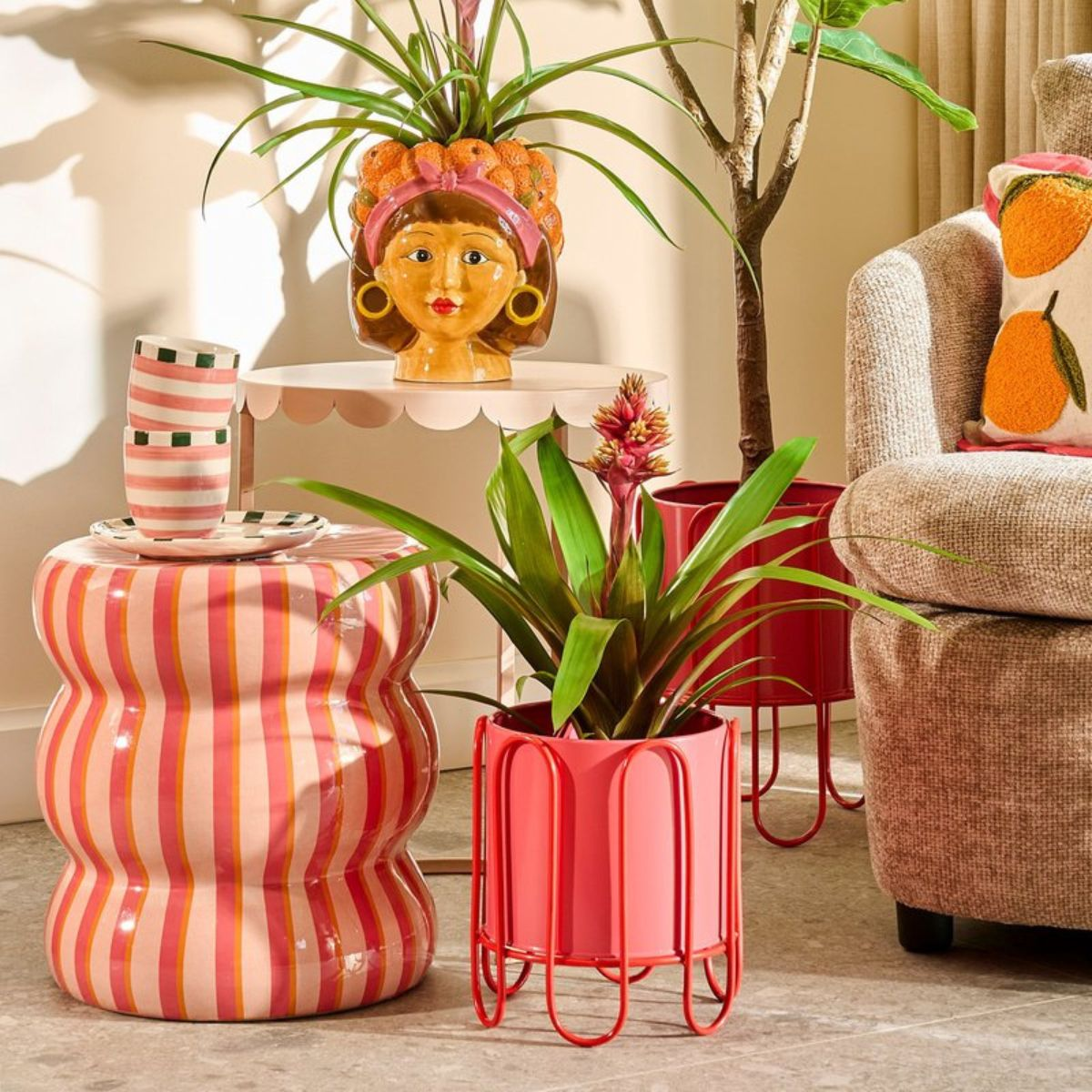Un salon coloré avec tabouret rayé rose, plantes en pot, vase tête et fauteuil avec coussins orange.