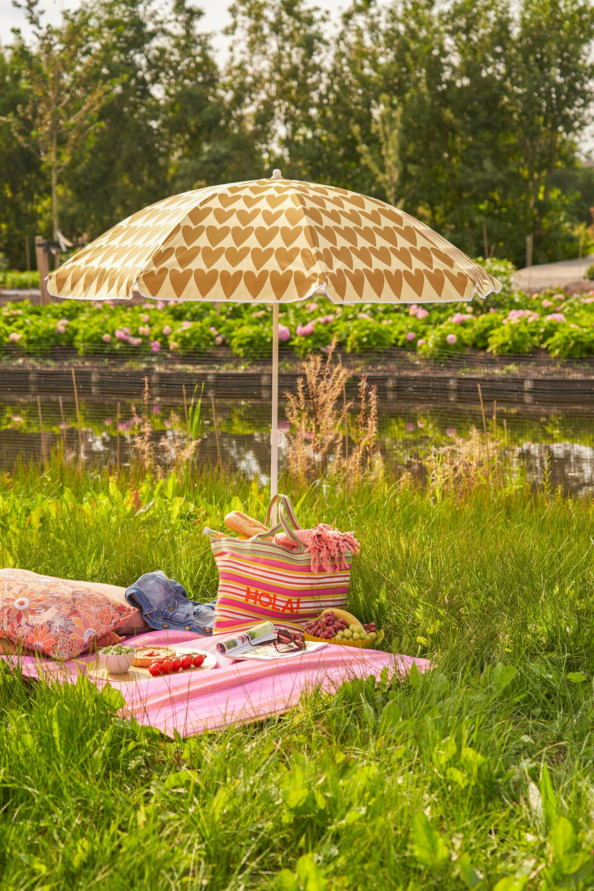 Eine pinke Picknickdecke mit herzgemustertem Schirm, gestreifter Tasche, Essen und Obst an einem Teich.