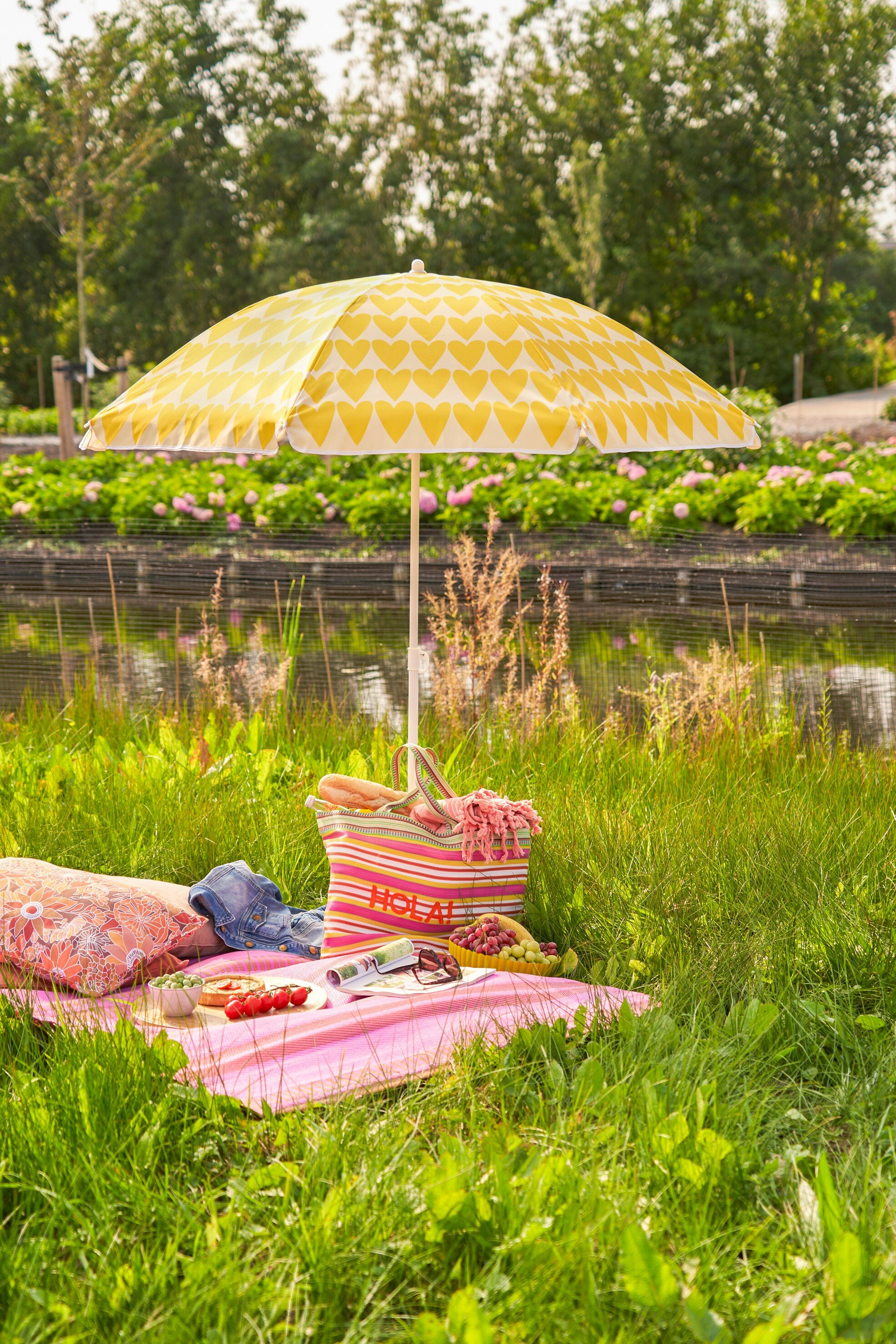 Ein gelb gemusterter Schirm spendet Schatten auf einer rosa Picknickdecke mit Essen, Tasche und Kissen im Gras.