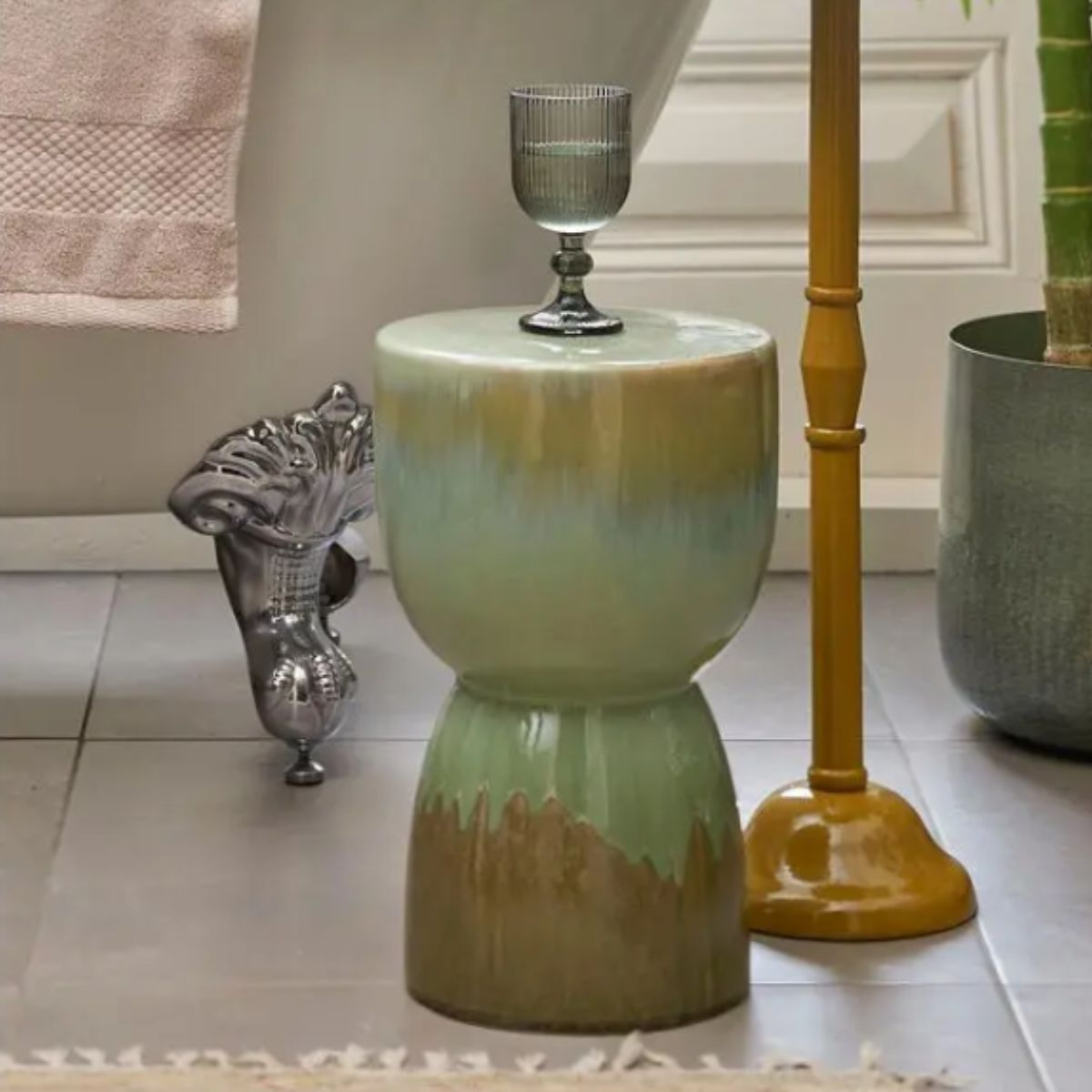 Ein grün-brauner Keramikhocker steht auf Fliesen, darauf ein graues Glas; links eine Badewanne, rechts eine gelbe Stange.