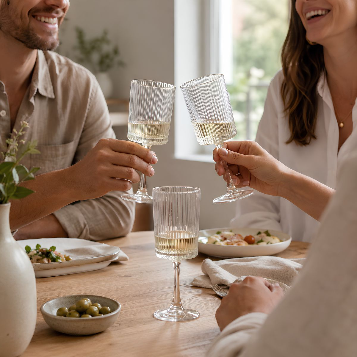 Menschen stoßen mit gerillten Weingläsern an einem hellen Holztisch mit Essen an.
