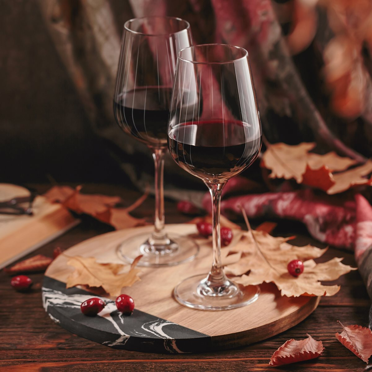 Zwei Gläser Rotwein auf einem Holzbrett, dekoriert mit Herbstblättern und roten Beeren.
