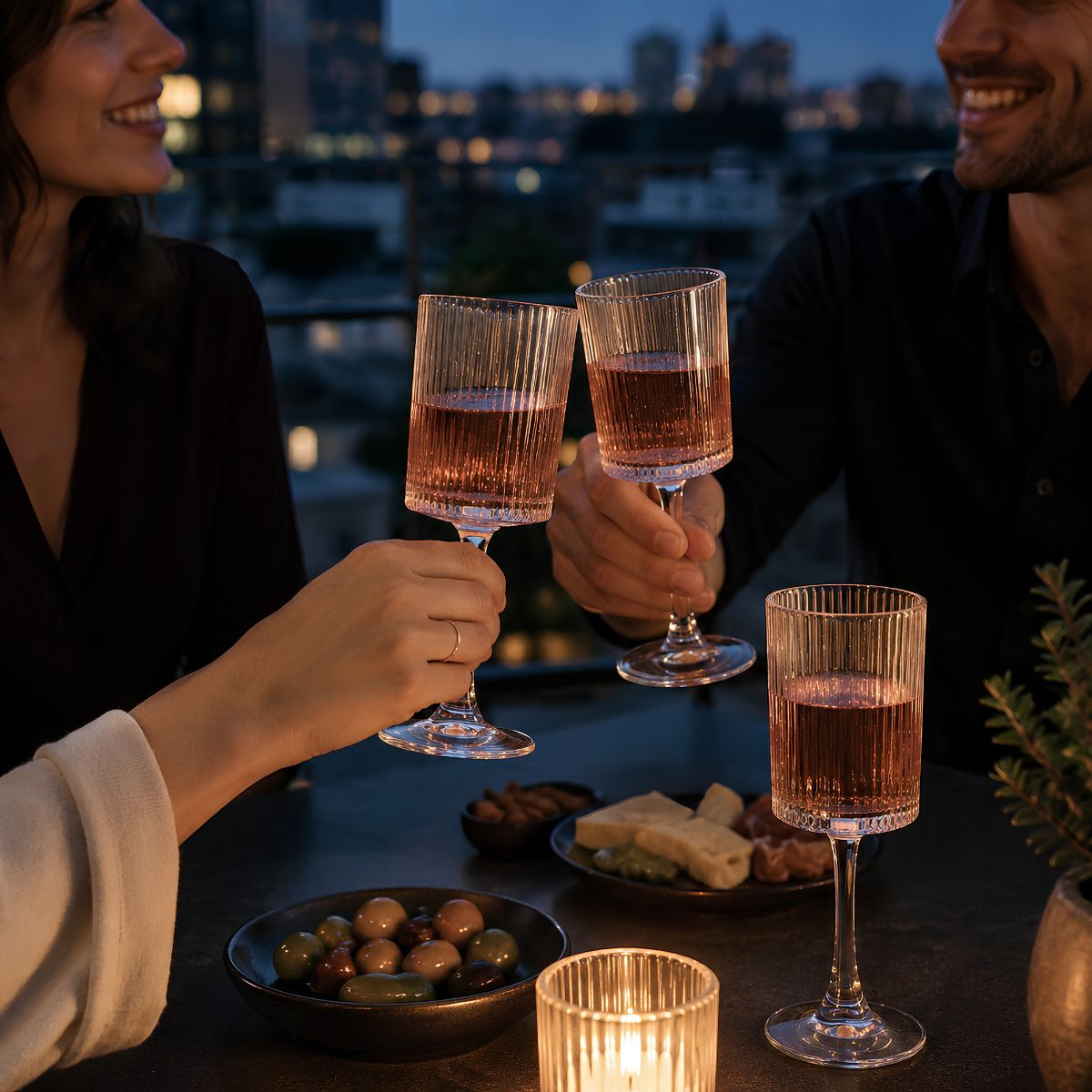 Paar stößt auf Dachterrasse mit Roséweingläsern bei Dämmerung an, mit Oliven, Käse und Kerzenschein.
