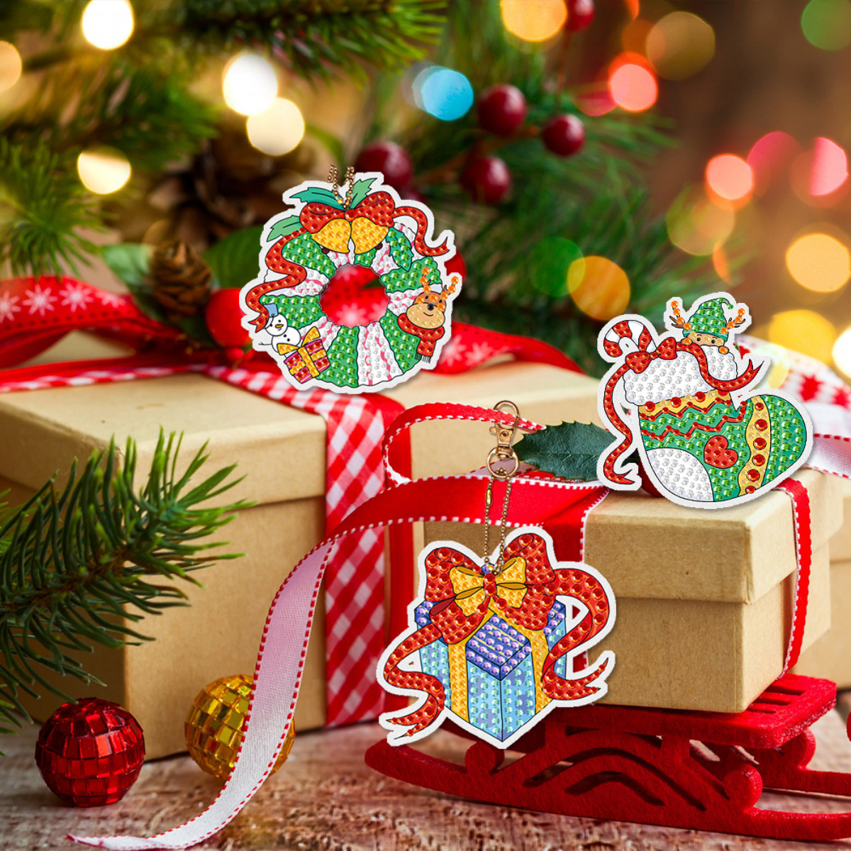 Drei diamantgemalte Weihnachtsschmuckstücke auf verpackten Geschenkkartons mit leuchtenden Lichtern im Hintergrund.