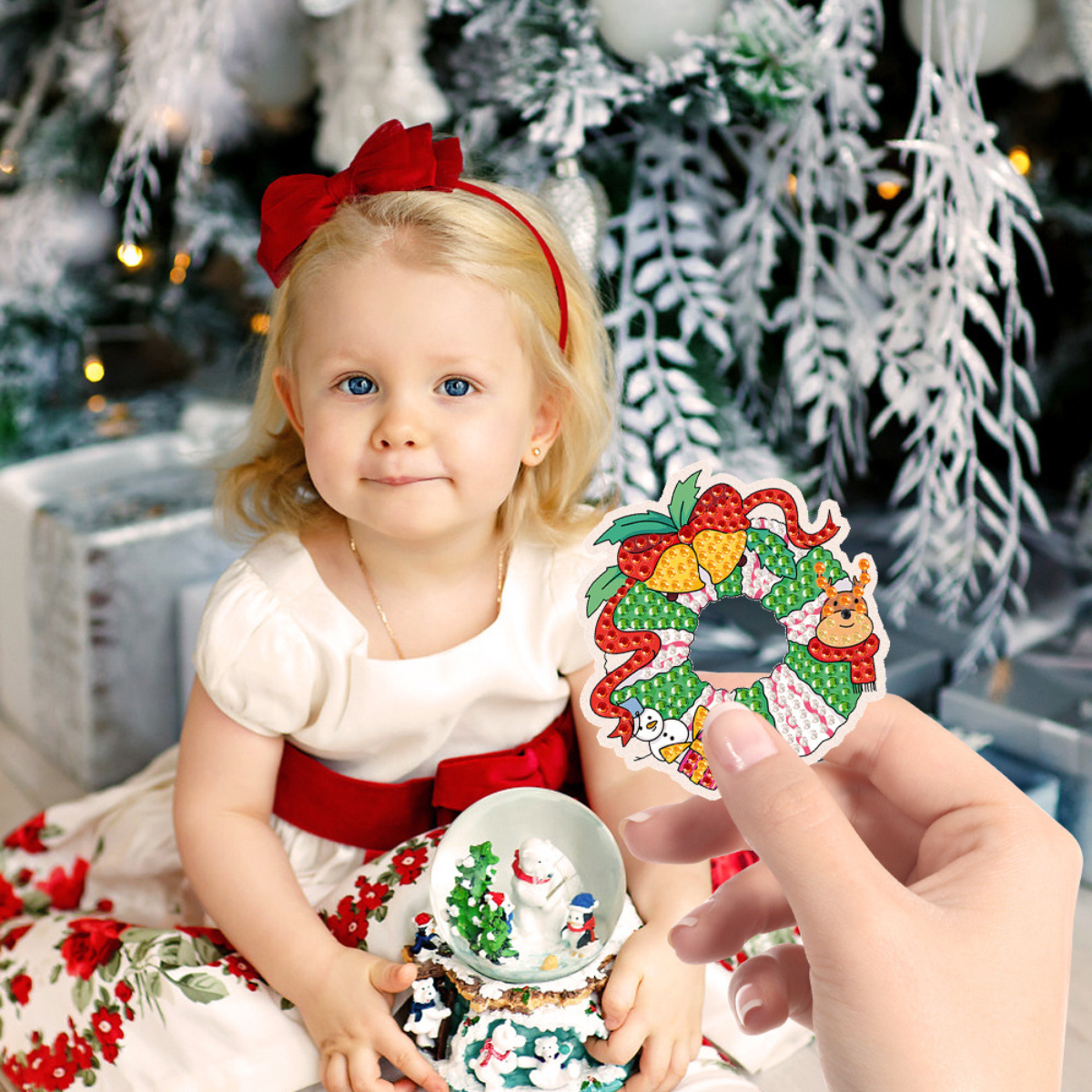 Ein blondes Mädchen in weißem Kleid mit roter Schleife hält eine Schneekugel, während eine Hand einen Weihnachtskranz zeigt.