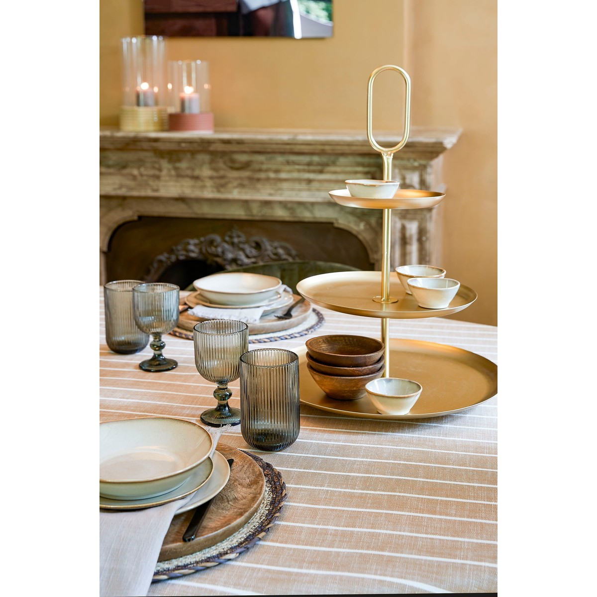 Ein gedeckter Tisch mit gestreifter Tischdecke, Tellern, gerippten Gläsern, Holzschalen und einer goldenen Etagère.