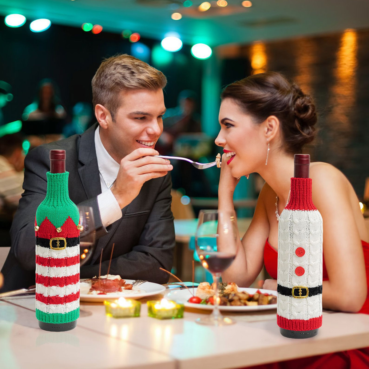 Ein Paar sitzt im Restaurant, der Mann füttert die Frau. Im Vordergrund stehen zwei Weinflaschen mit Weihnachtspullovern.