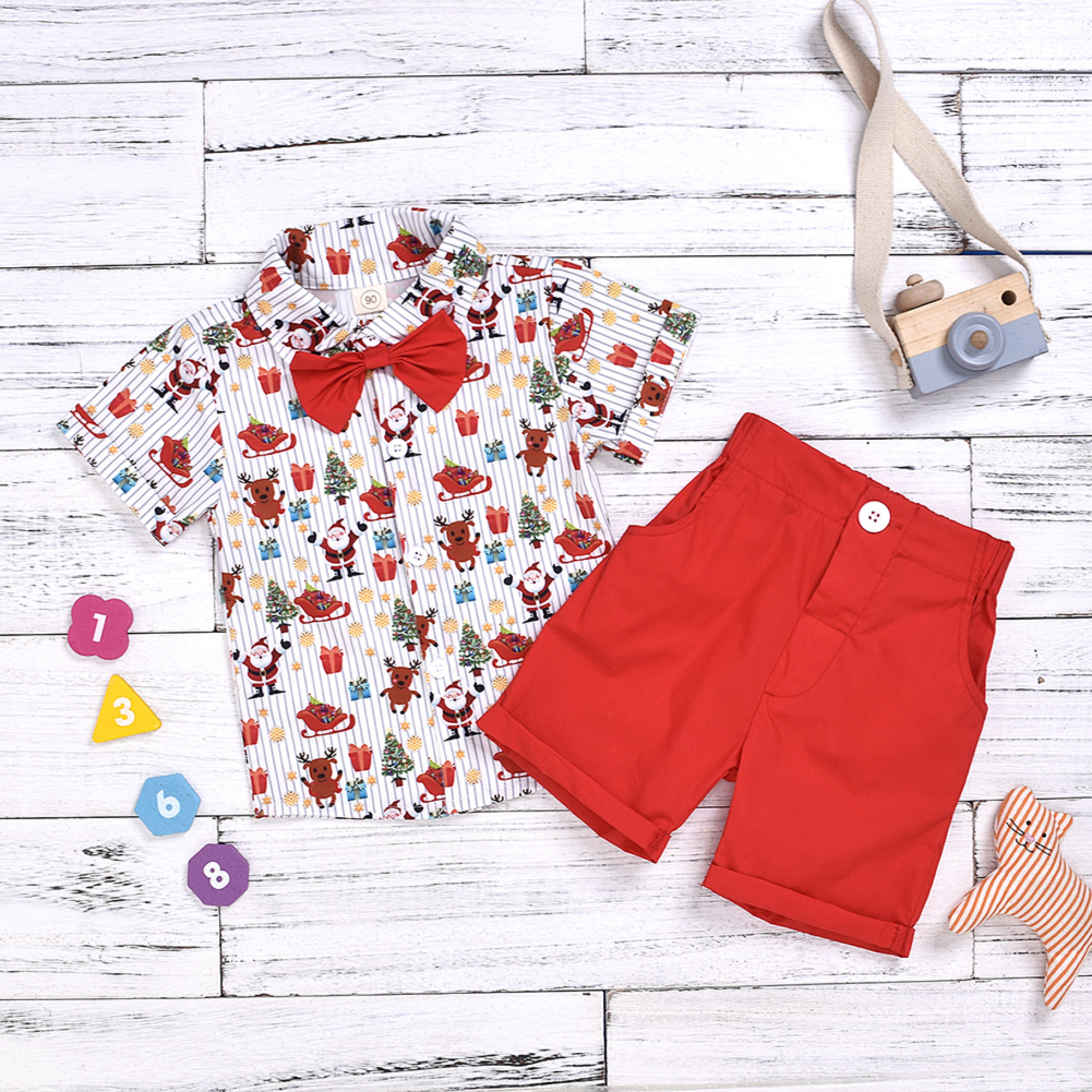 Weihnachtsoutfit für Jungen mit gemustertem Hemd, roter Fliege und roten Shorts auf Holzgrund.