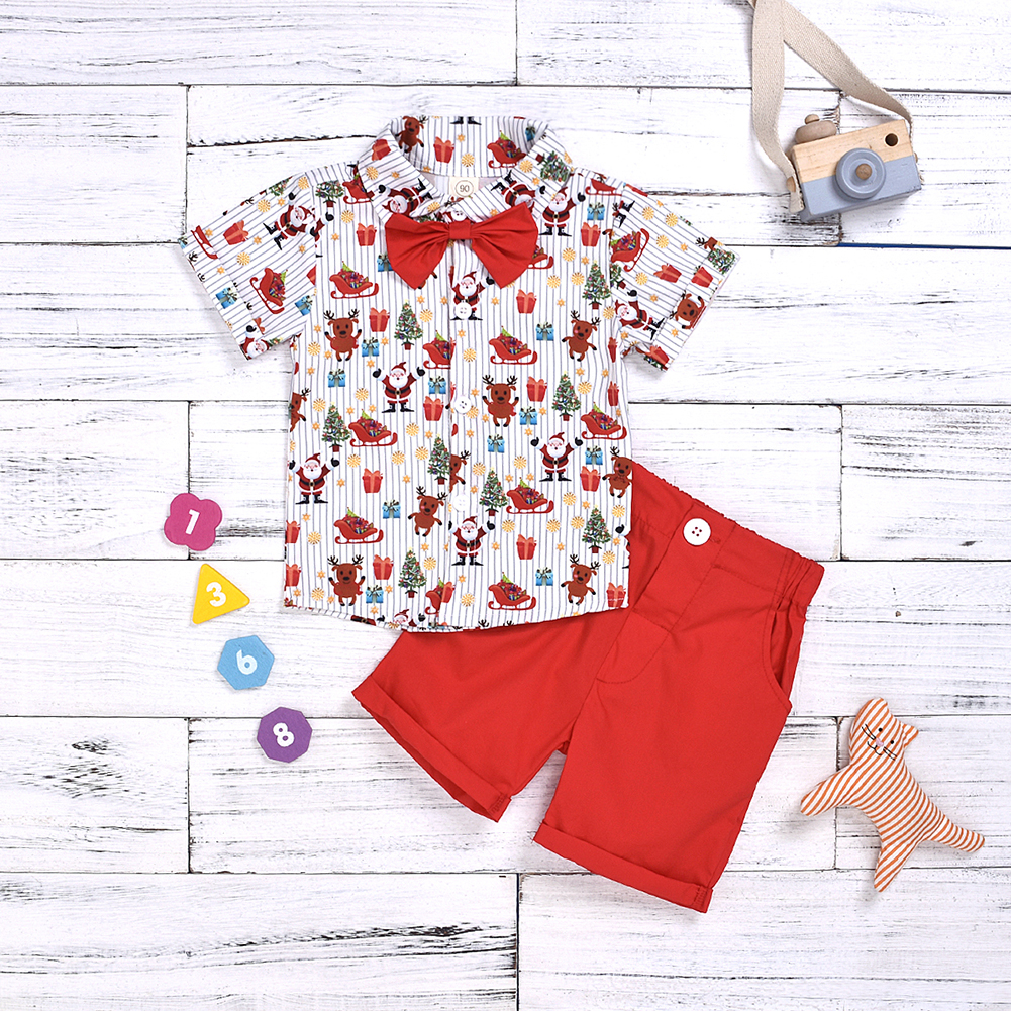 Ein Kinderweihnachtsoutfit mit gemustertem Hemd, roter Fliege und roten Shorts auf weißem Holzhintergrund.