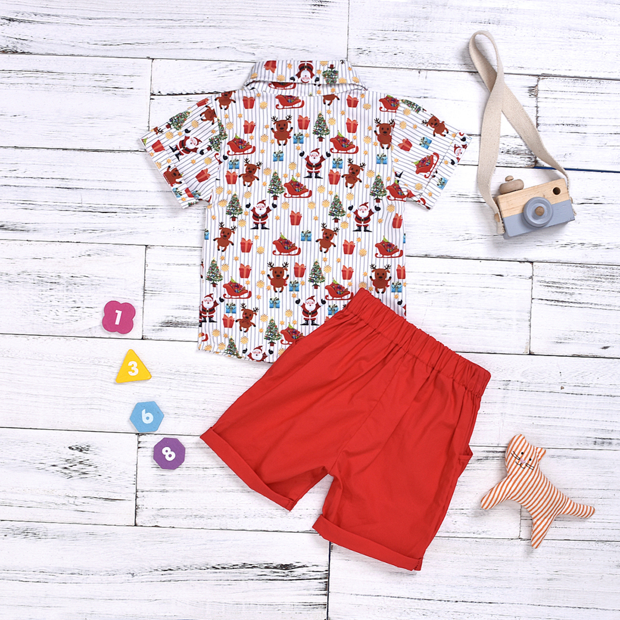 Ein weihnachtliches Kinderoutfit mit gemustertem Hemd und roten Shorts auf weißem Holzhintergrund.