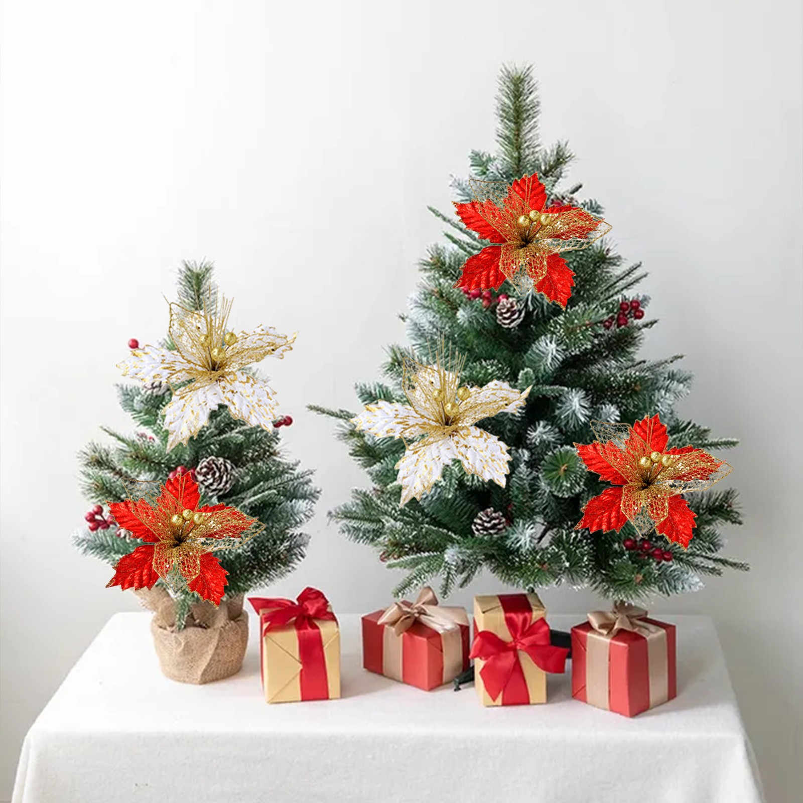 Zwei kleine Weihnachtsbaeume mit roten und goldenen Blumen geschmueckt, umgeben von Geschenken auf weissem Tisch.