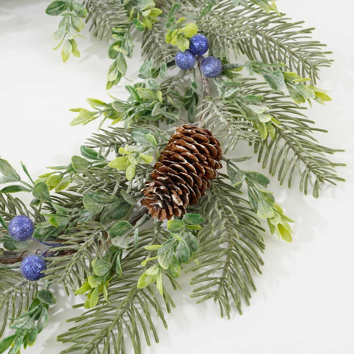 Eine winterliche Girlande mit Tannenzapfen, grünen Nadeln und glitzernden blauen Beeren auf weißem Hintergrund.