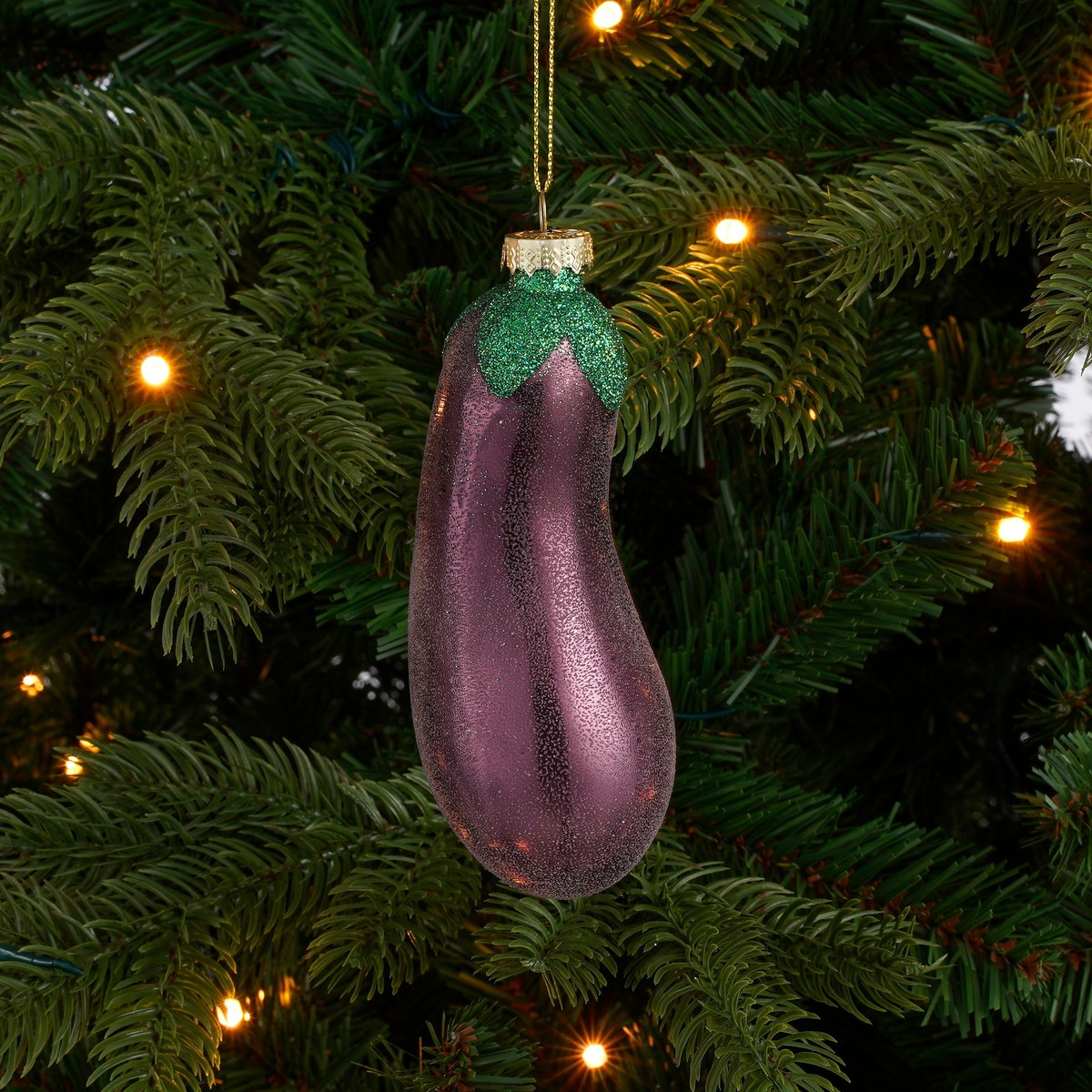 Glänzende, purpurne Auberginen-Christbaumkugel hängt an einem Weihnachtsbaum mit Lichtern.