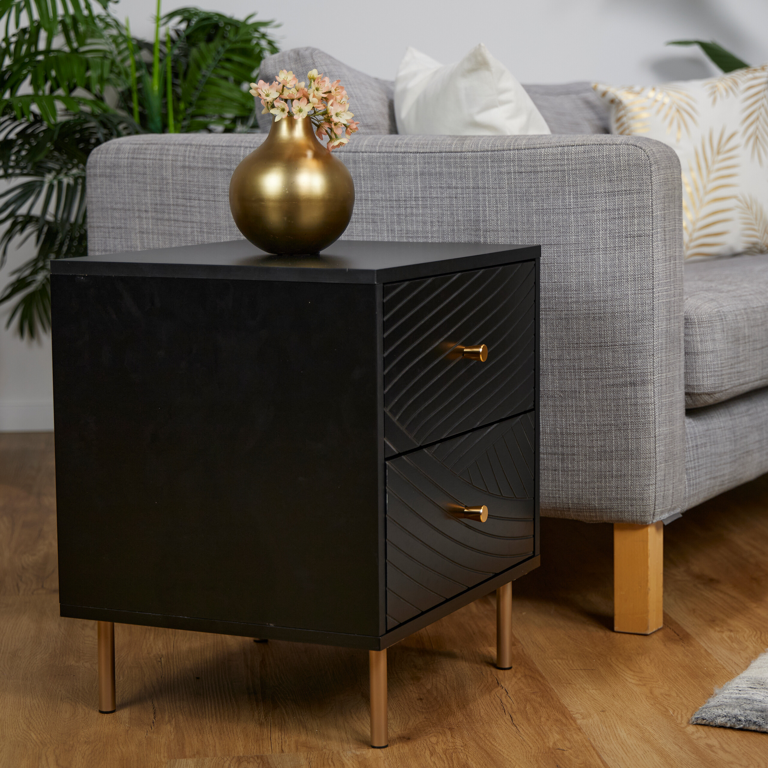 Ein schwarzer Nachttisch mit goldenen Beinen und einer goldener Vase mit Blumen darauf in einem Wohnzimmer mit grauem Sofa.